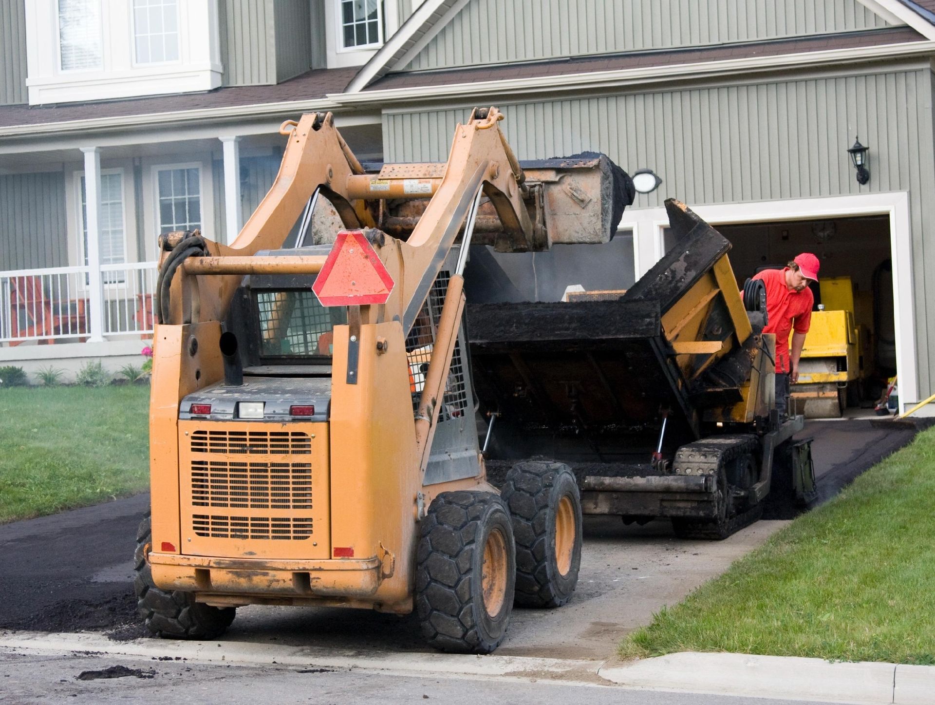 A worker in a red shirt operates a yellow skid-steer loader to spread asphalt on a residential driveway.