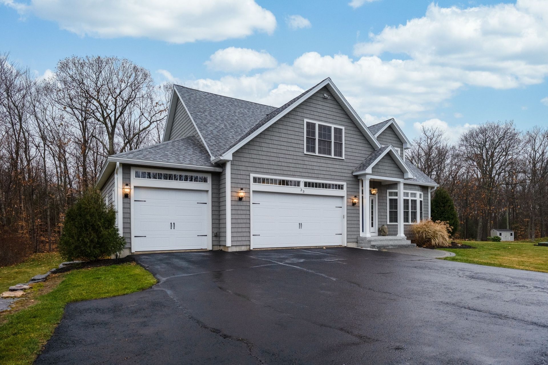 A gray, two-story house with a three-car garage and a dark asphalt driveway, set against a backdrop of bare trees.