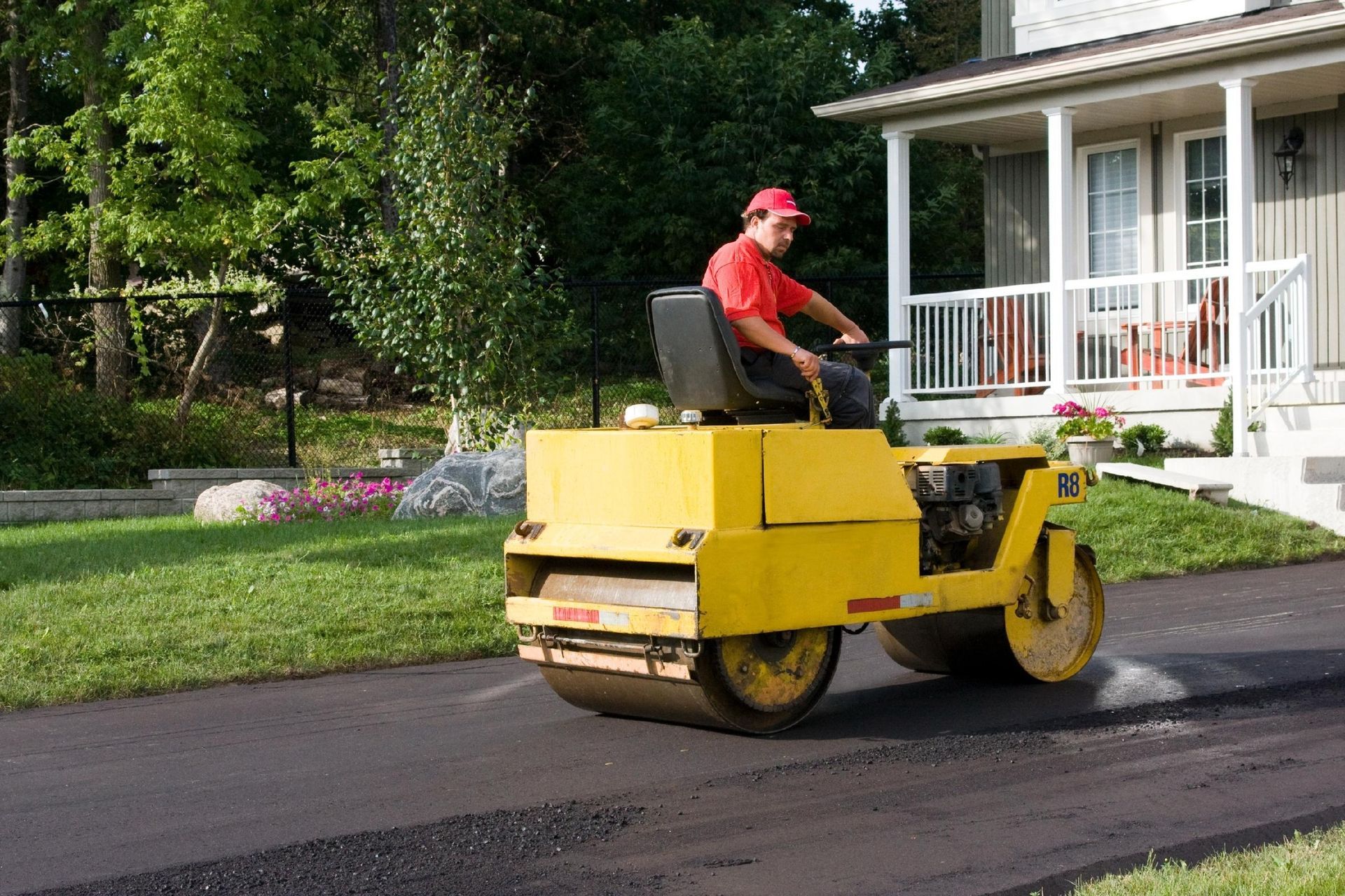 A person in a red shirt operates a yellow steamroller to pave a dark asphalt driveway in front of a house.