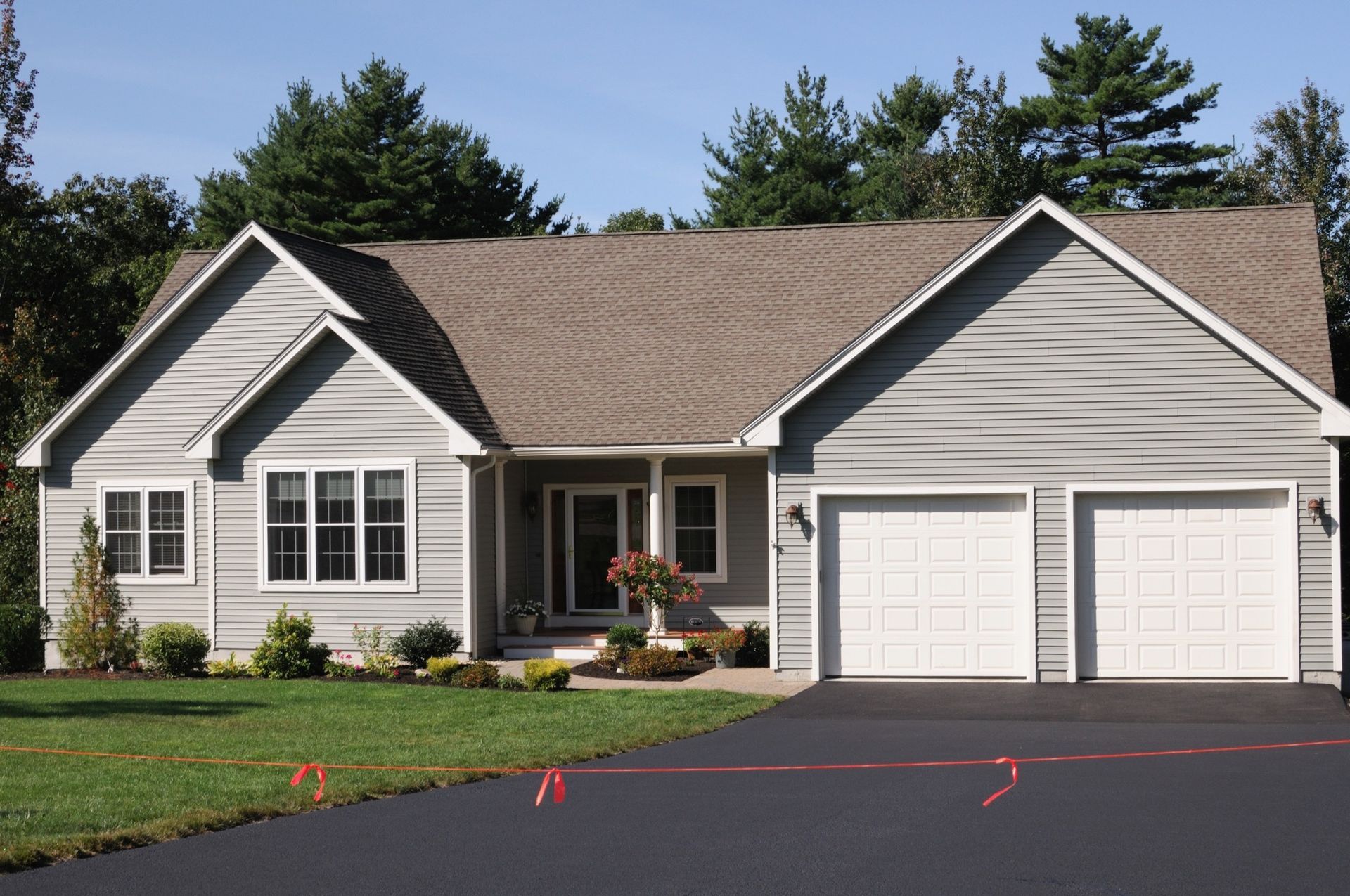A light gray house with two garage doors, a shingled roof, and a paved driveway surrounded by grass and trees.