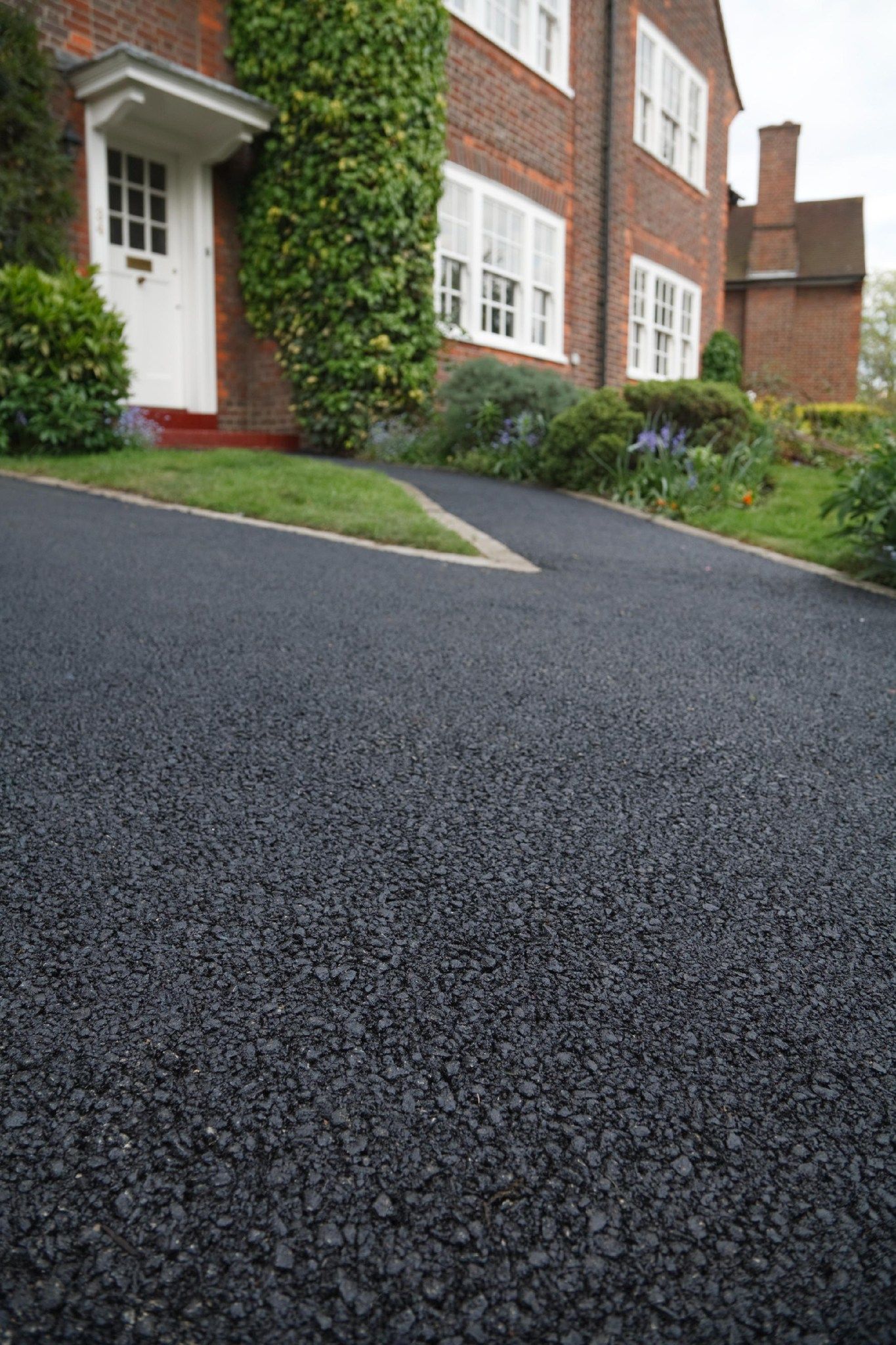 A freshly paved black asphalt driveway leading to the white front door of a brick house surrounded by greenery.