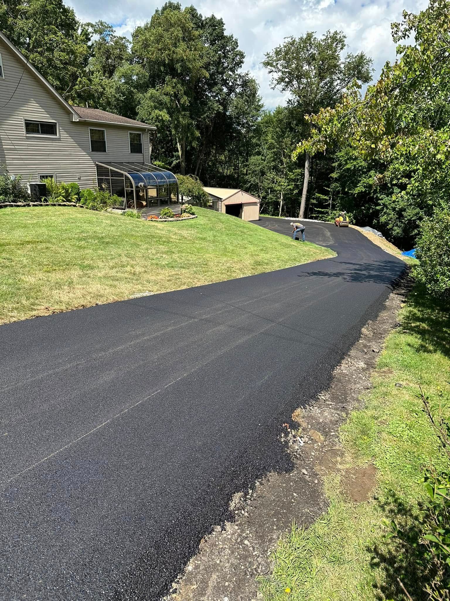 A freshly paved asphalt driveway curves past a residential house and trees toward a detached garage on a sunny day.