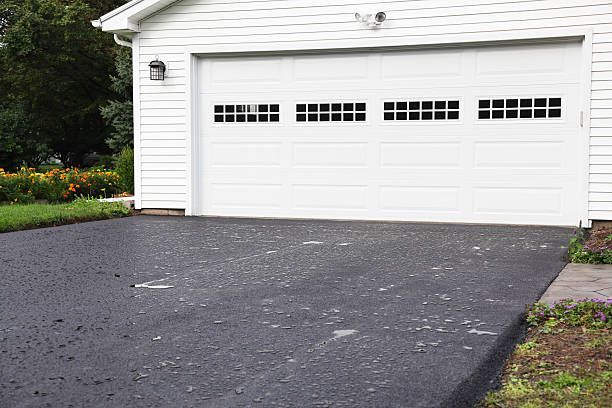 A white garage door with rectangular windows above a freshly paved black asphalt driveway.