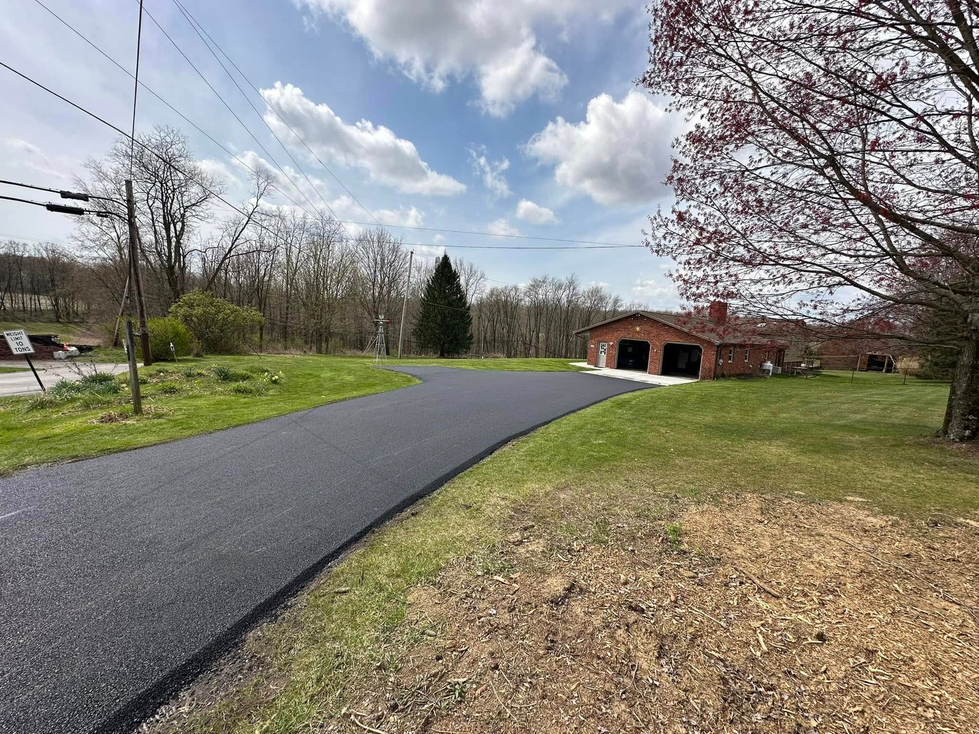 A freshly paved asphalt driveway leads to a brick garage in a grassy, suburban landscape under a partly cloudy sky.