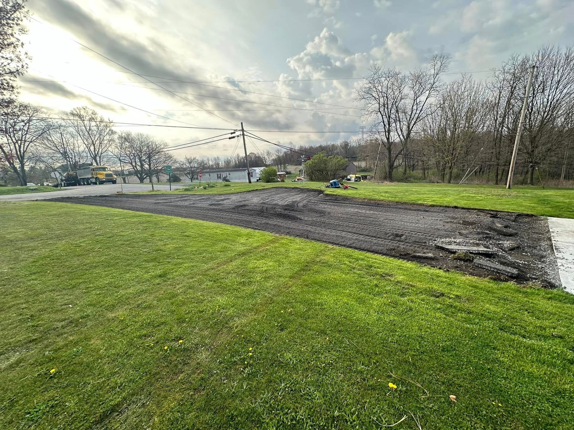 A rectangular patch of dark, tilled soil sits in a green lawn under a cloudy sky near a line of bare trees.