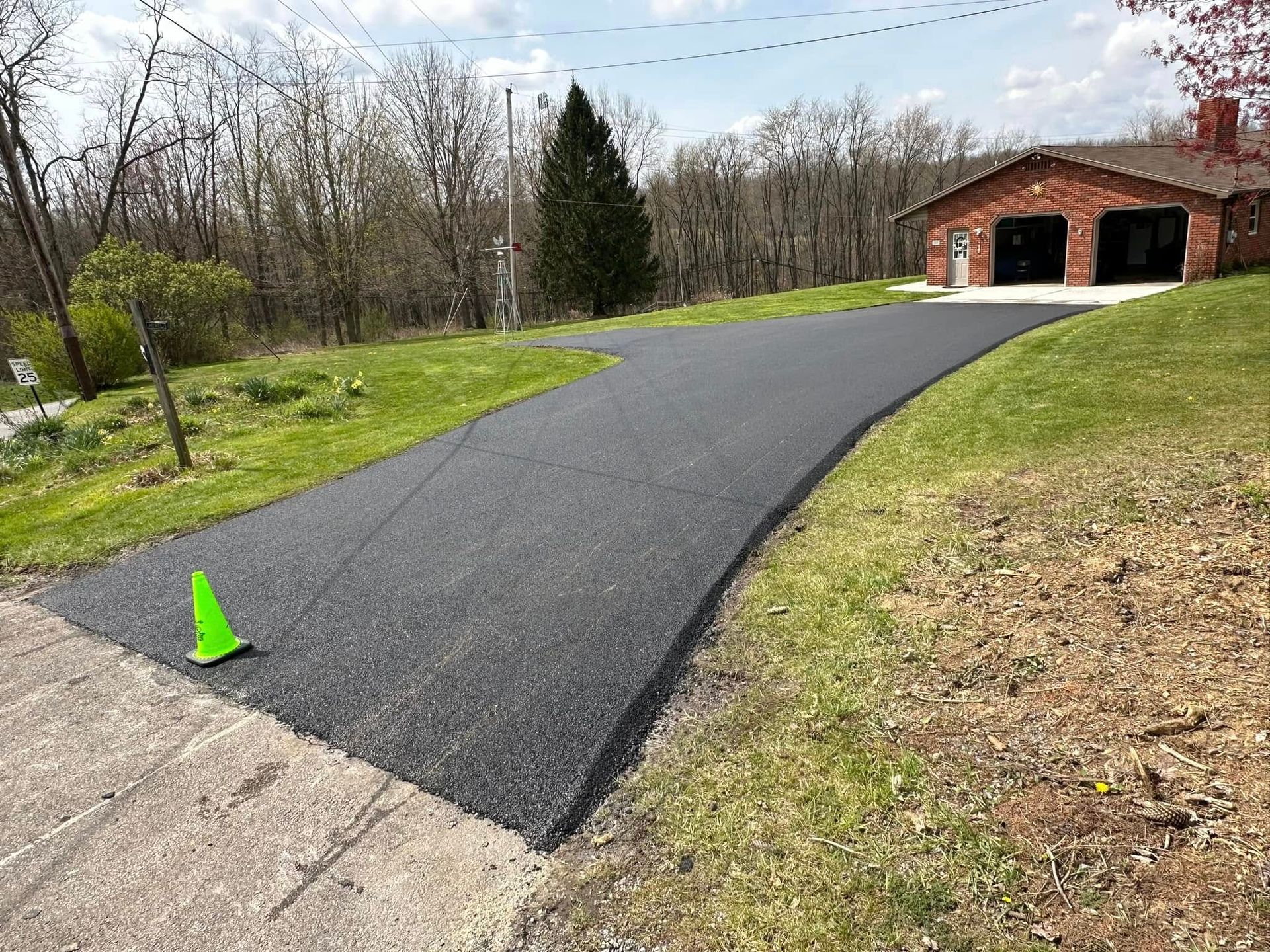 Freshly paved asphalt driveway leading to a brick two-car garage, with a lime green cone on the lower left corner.