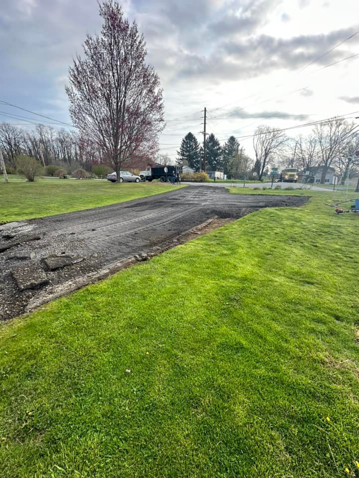 A section of dirt and gravel driveway under construction, surrounded by green grass with a tree and road in the background.