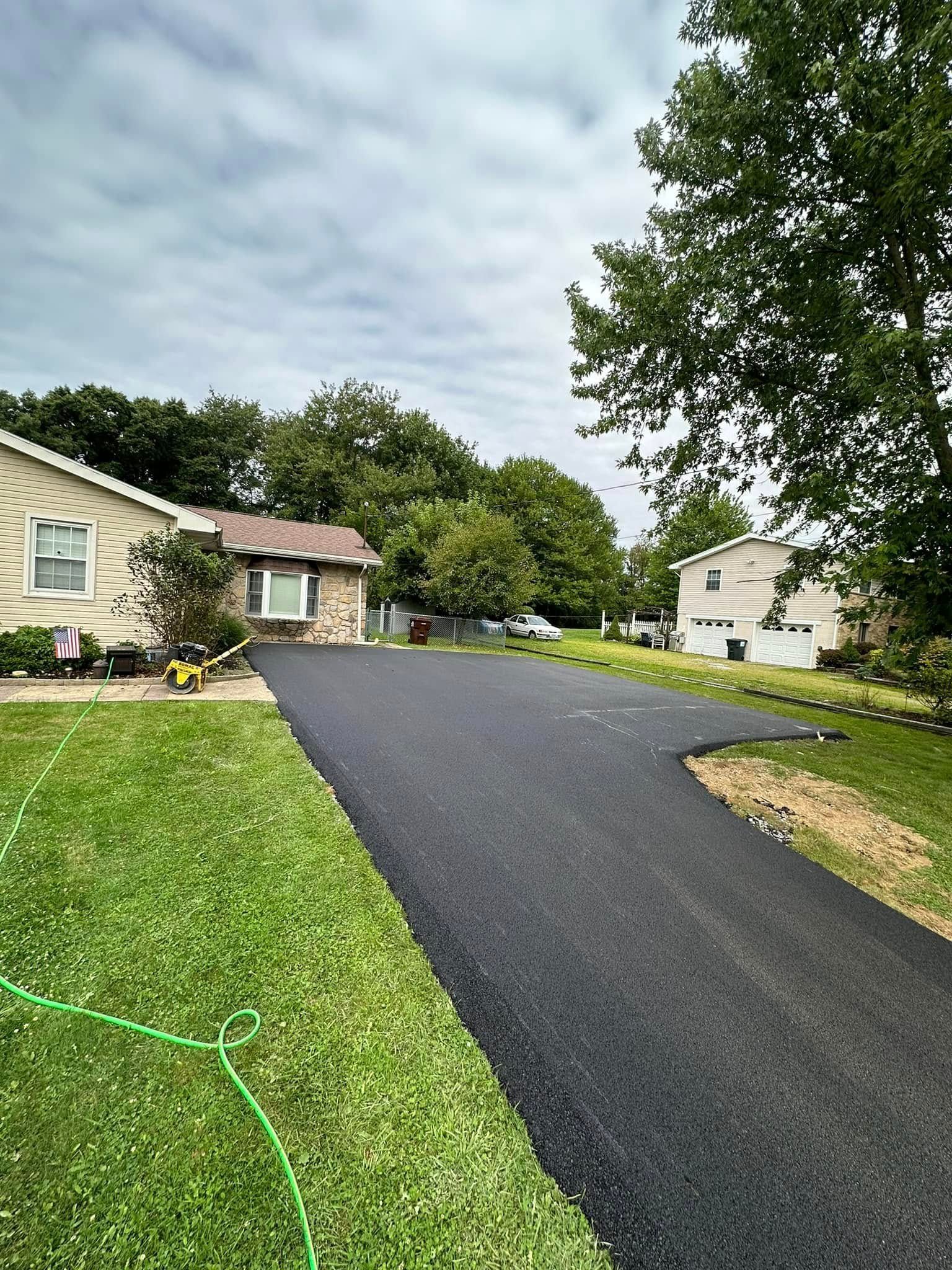 A newly paved black asphalt driveway leads to a light-colored house with green grass and trees under a cloudy sky.