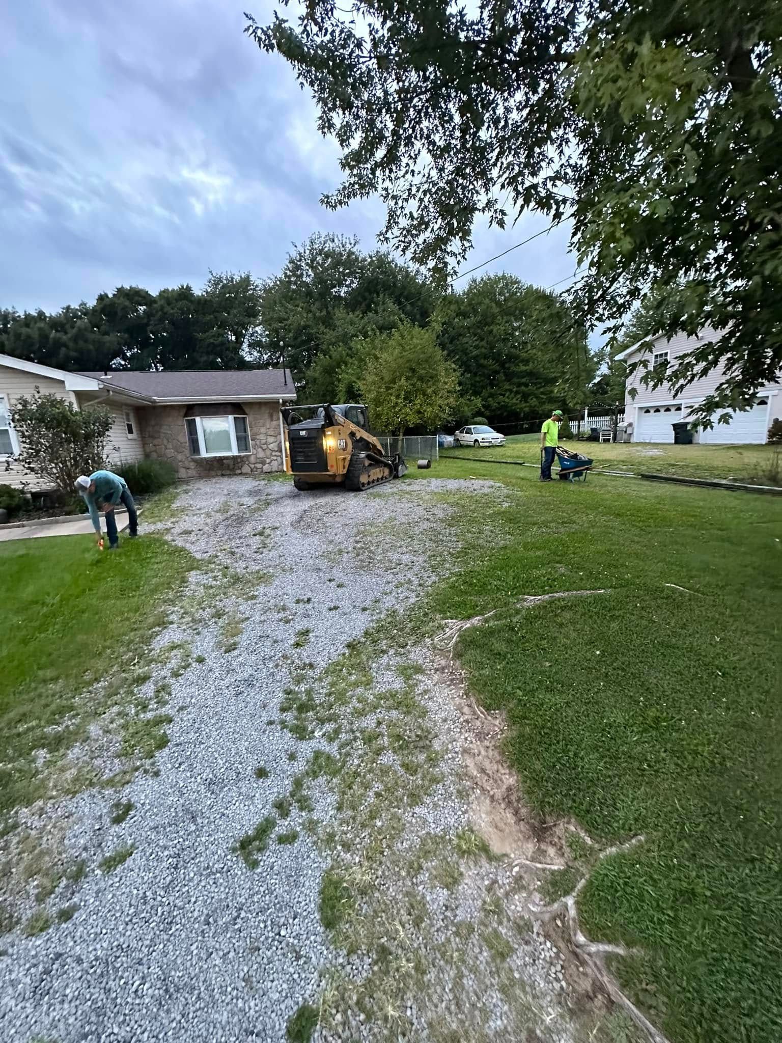 Workers use a yellow skid steer to grade a gravel driveway in front of a house on a cloudy day.