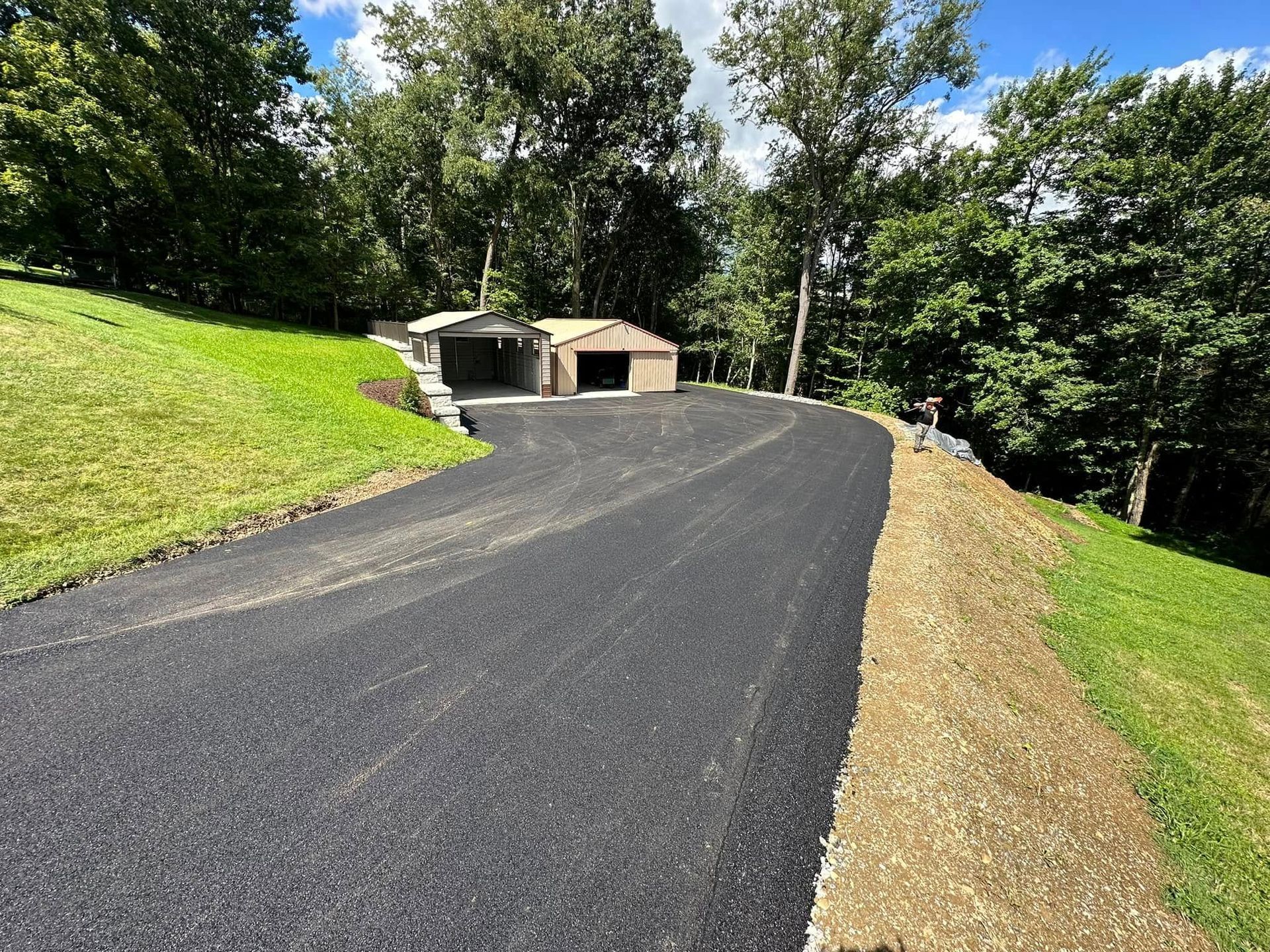 A paved asphalt driveway leads up to a tan detached garage surrounded by trees on a bright day.