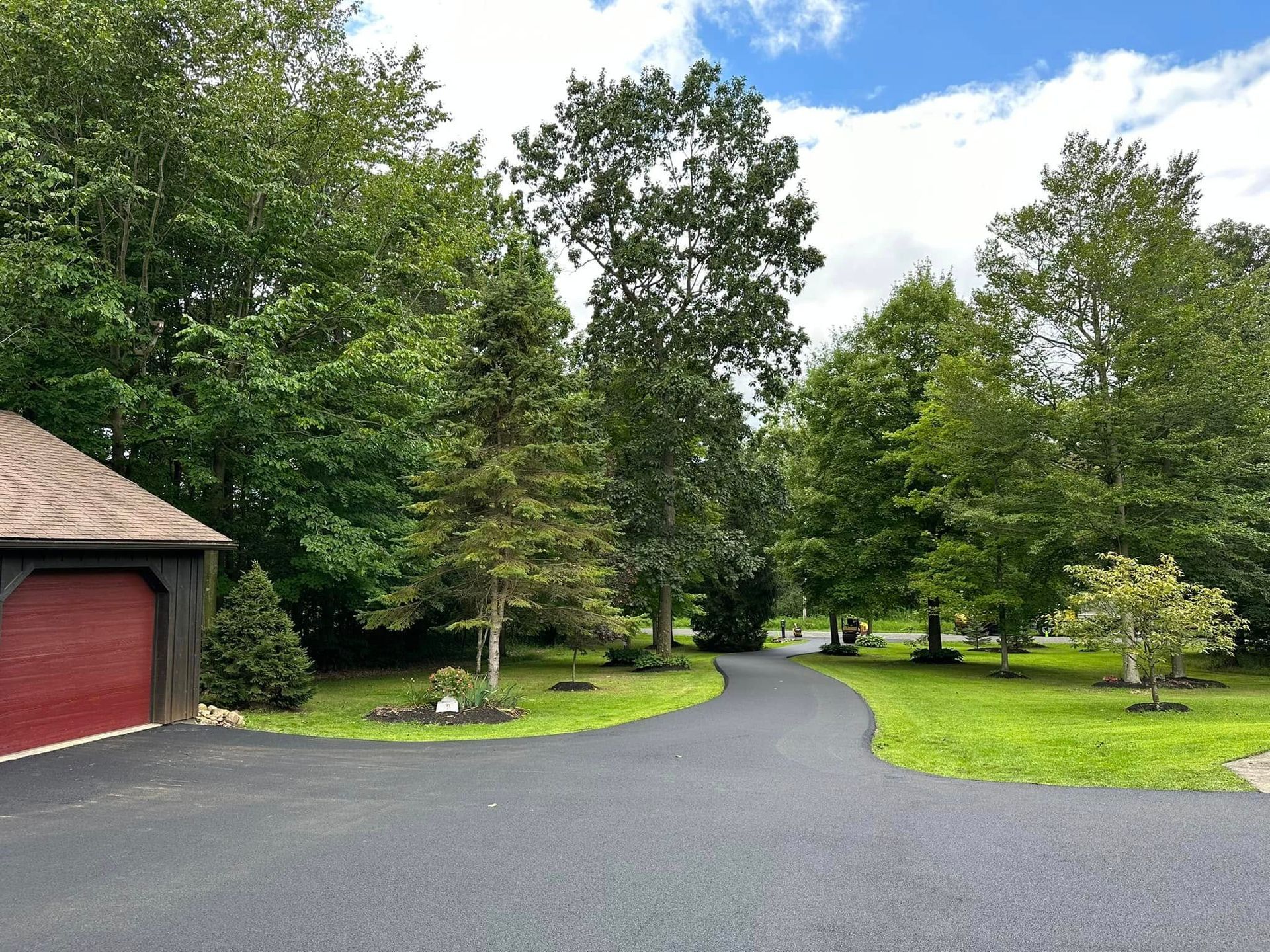 A paved driveway curves past a garage with a red door toward a house obscured by dense trees and green lawn.