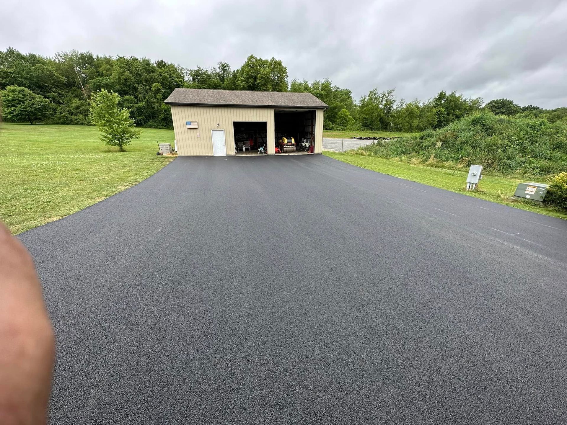 A newly paved, dark asphalt driveway leading to a tan detached garage on a grassy, overcast day.