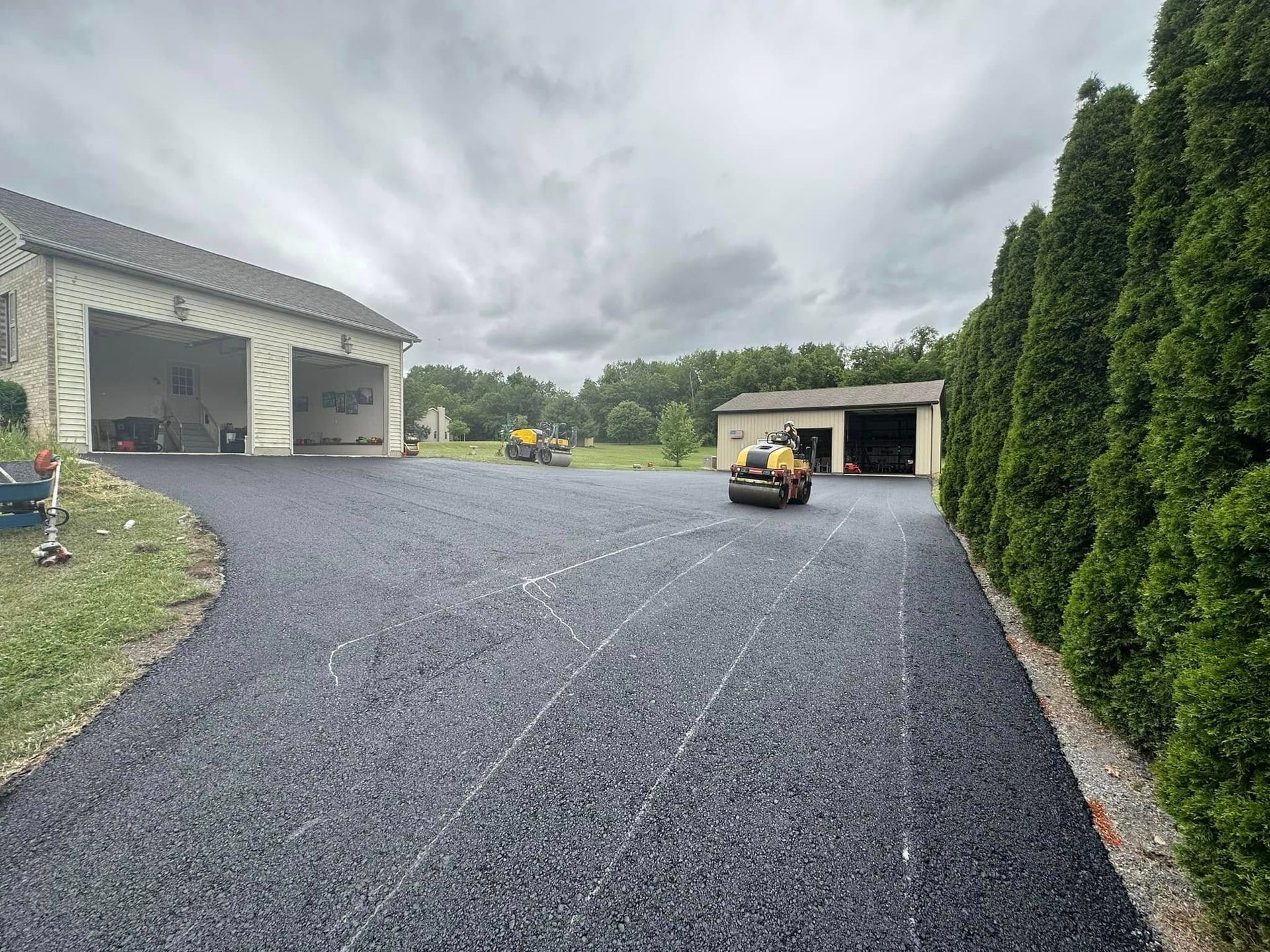 A freshly paved asphalt driveway connects two garage buildings on a cloudy day, with a small yellow roller parked nearby.