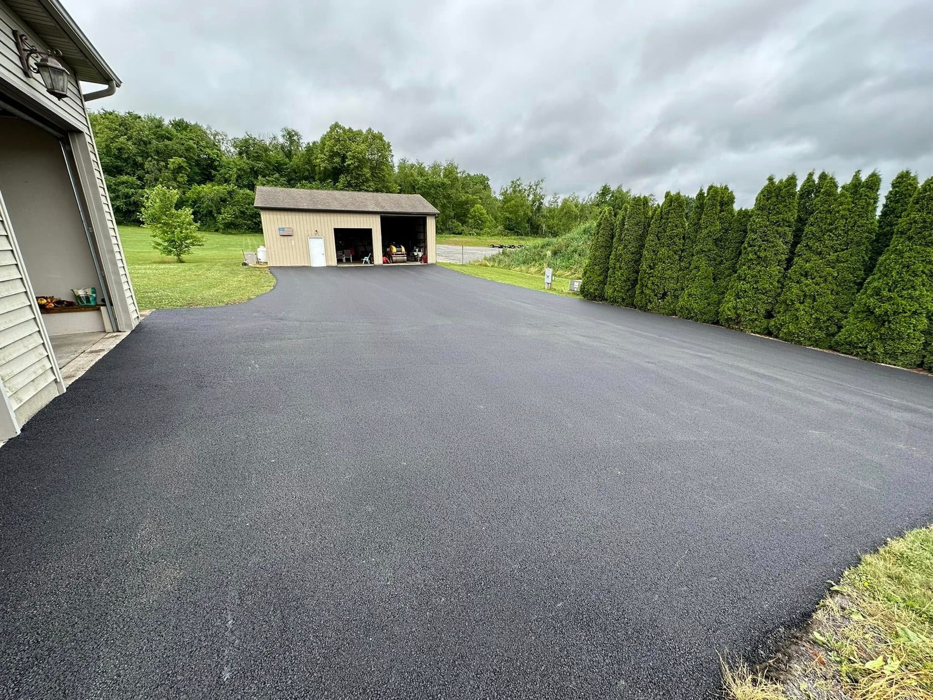A freshly paved dark asphalt driveway leads from the foreground toward a detached garage, flanked by a row of tall hedges.