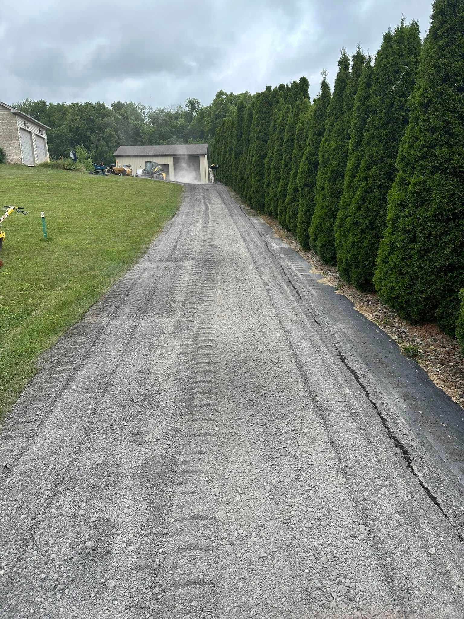 A gravel driveway lined by tall green evergreen trees leads to a garage in the distance under a cloudy sky.