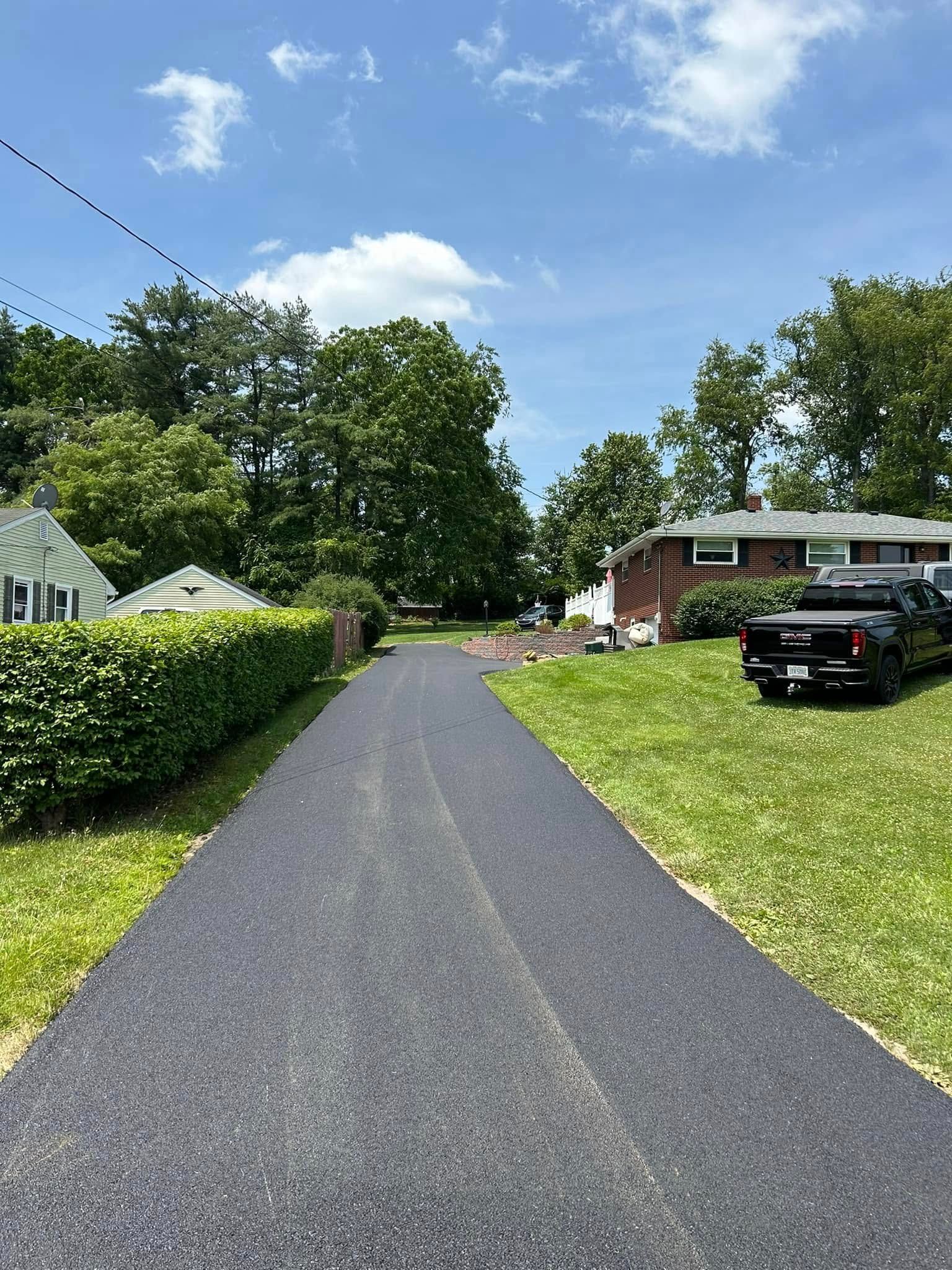 A newly paved asphalt driveway leads between a manicured green hedge and a grassy lawn toward a brick house.