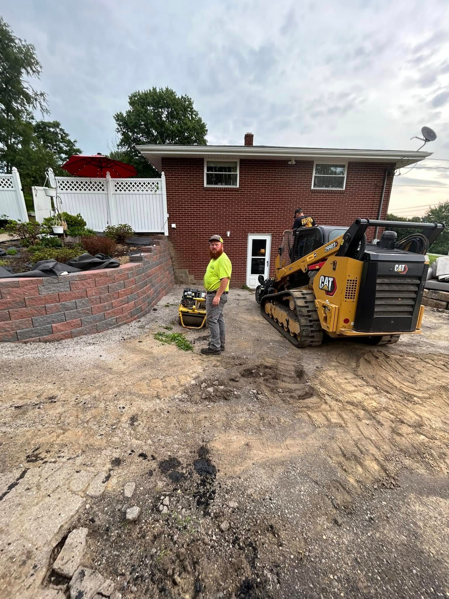 A person in a high-visibility vest stands near a yellow compact track loader in front of a house under renovation.