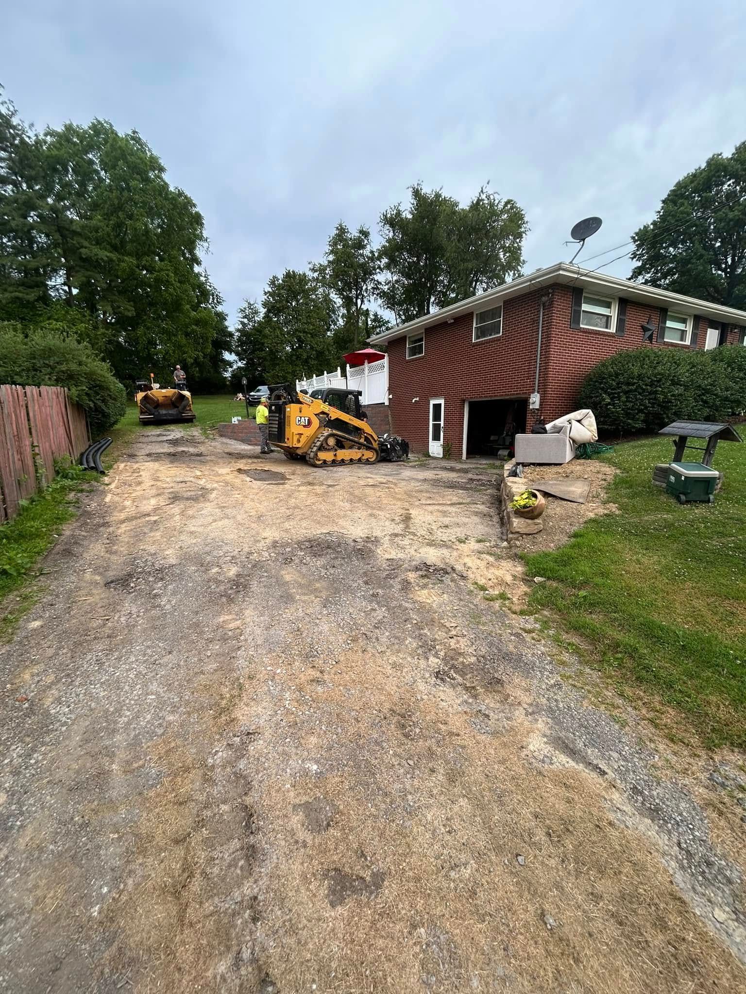 A yellow skid steer loader sits on a gravel driveway in front of a brick house during landscaping work.