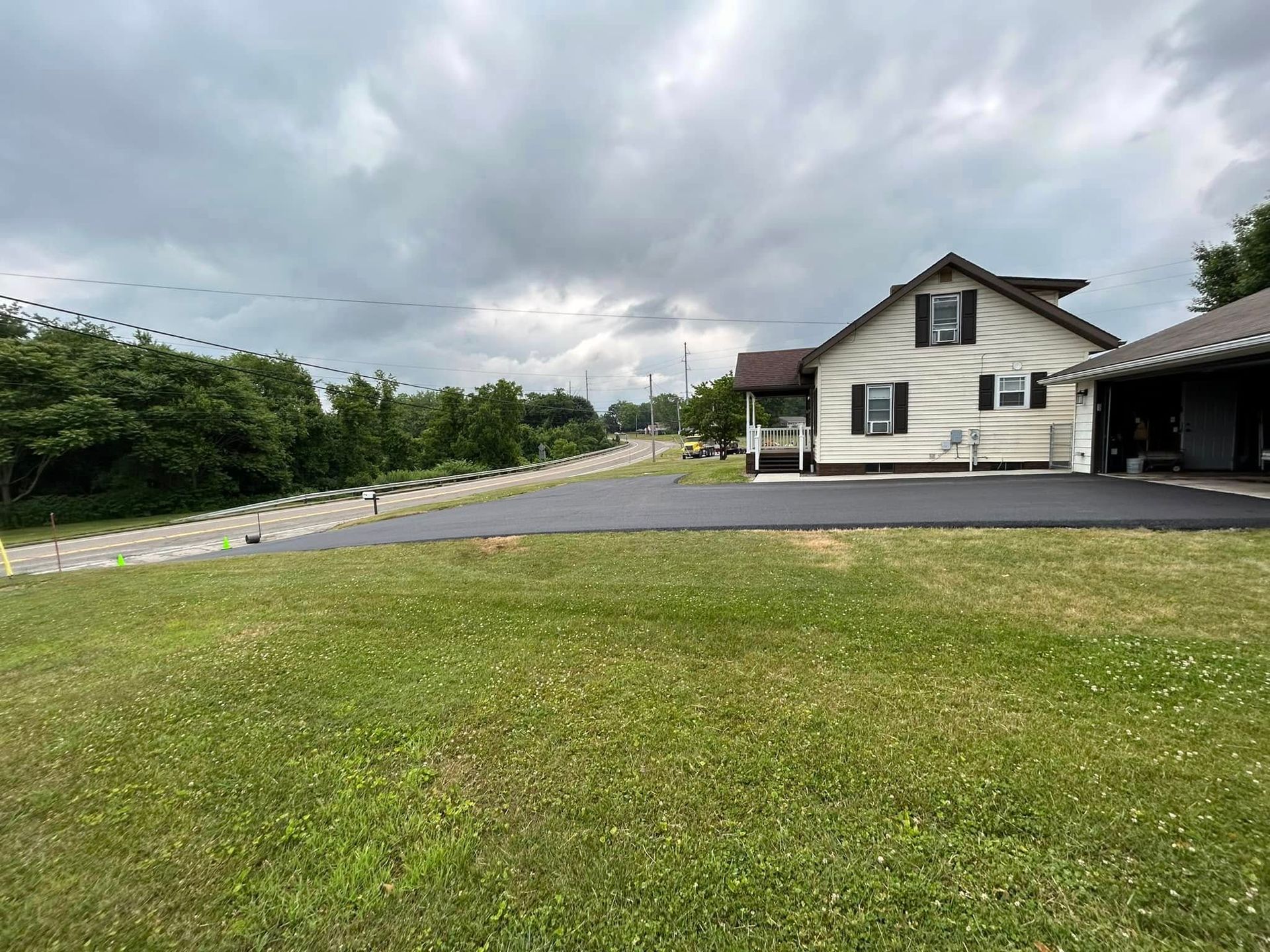 A white house with a new asphalt driveway and attached garage sits next to a lush grassy lawn under a cloudy sky.