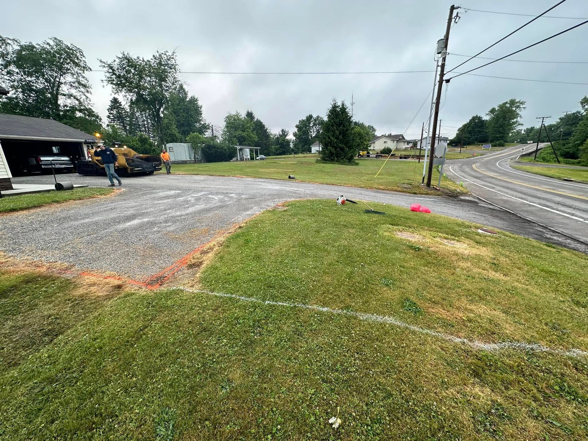 A gravel driveway leads from a road toward a dark garage, with two people working on the property on a cloudy day.