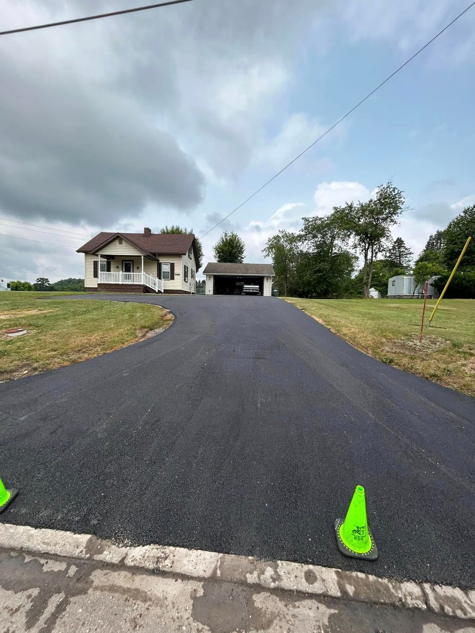 A newly paved asphalt driveway leads up a gentle slope toward a house and detached garage under a cloudy sky.
