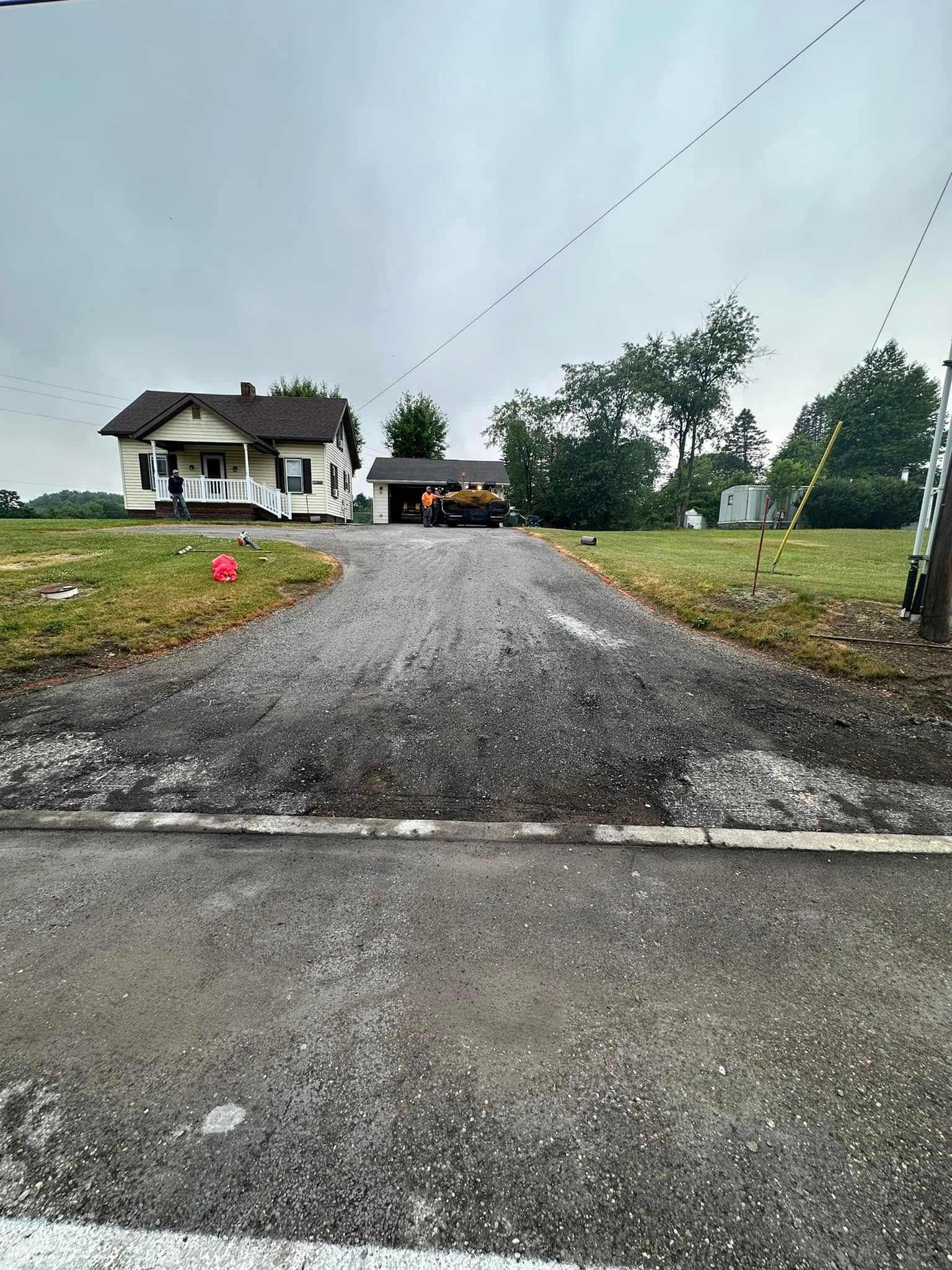 A gravel driveway leads from an asphalt road to a white house with a dark roof and a detached garage under a cloudy sky.