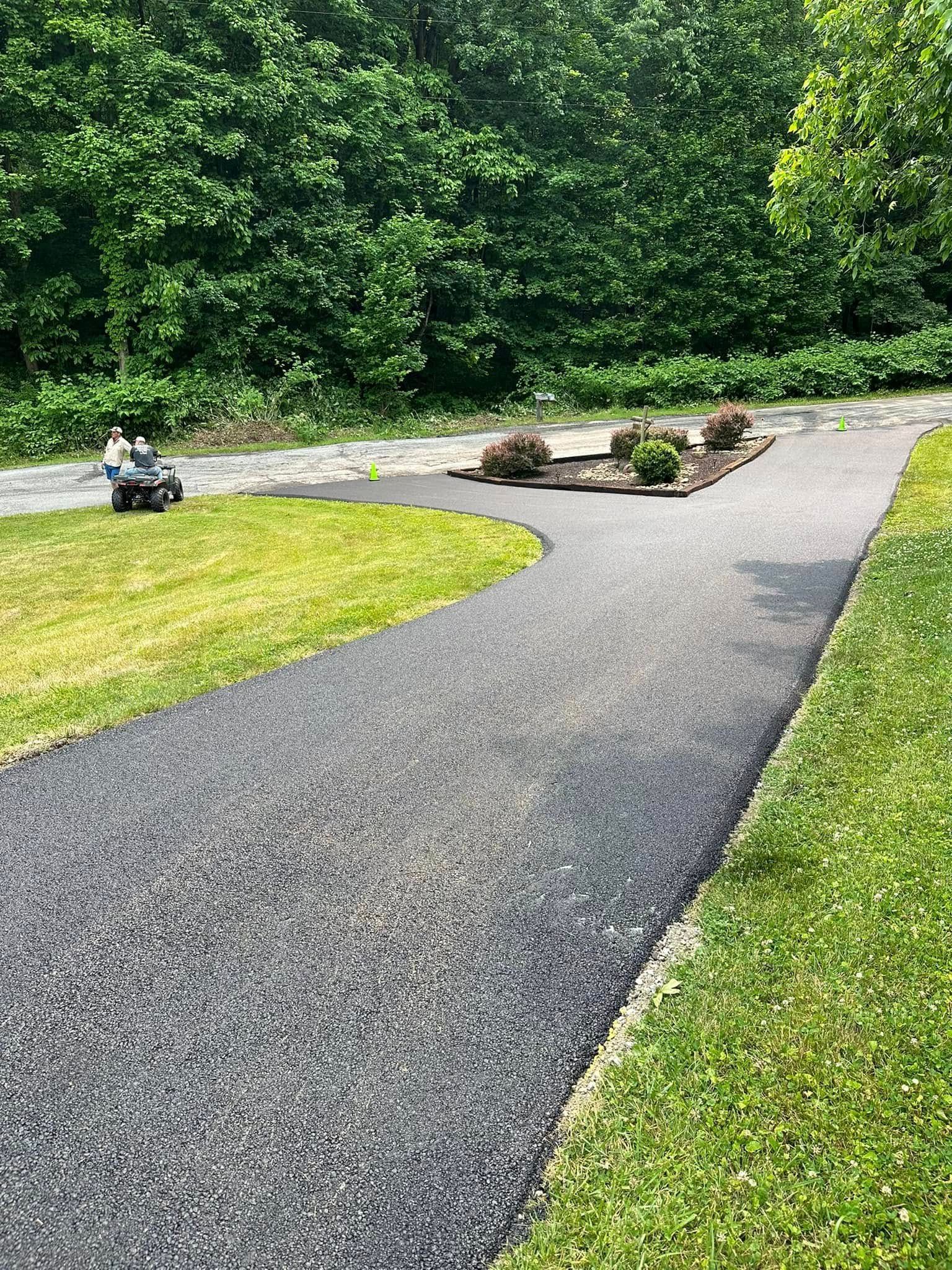 A freshly paved asphalt driveway curves through a green lawn, leading toward a garden bed with small shrubs and trees.