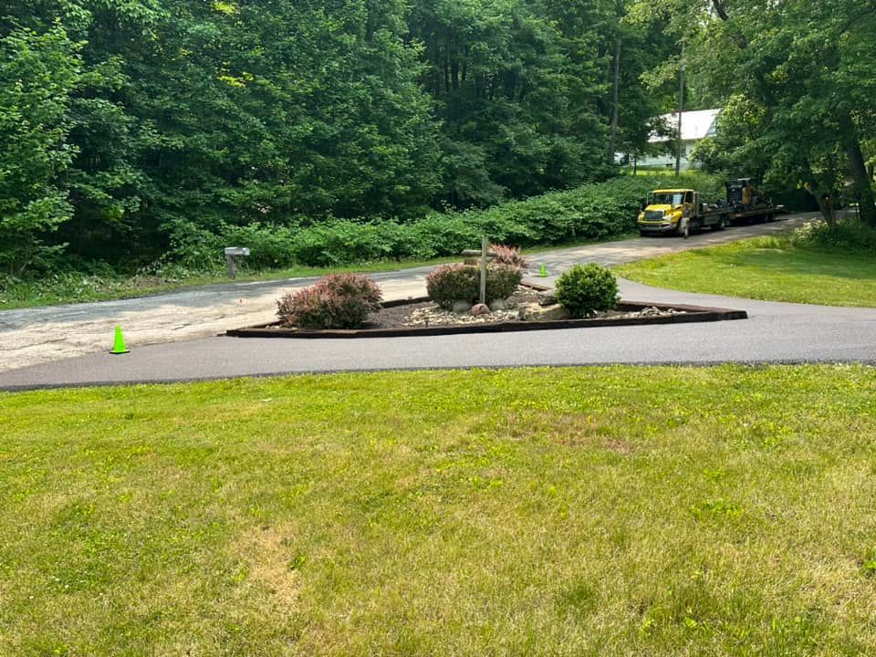 A triangular garden island sits in the center of a paved driveway, with a construction truck parked in the background.