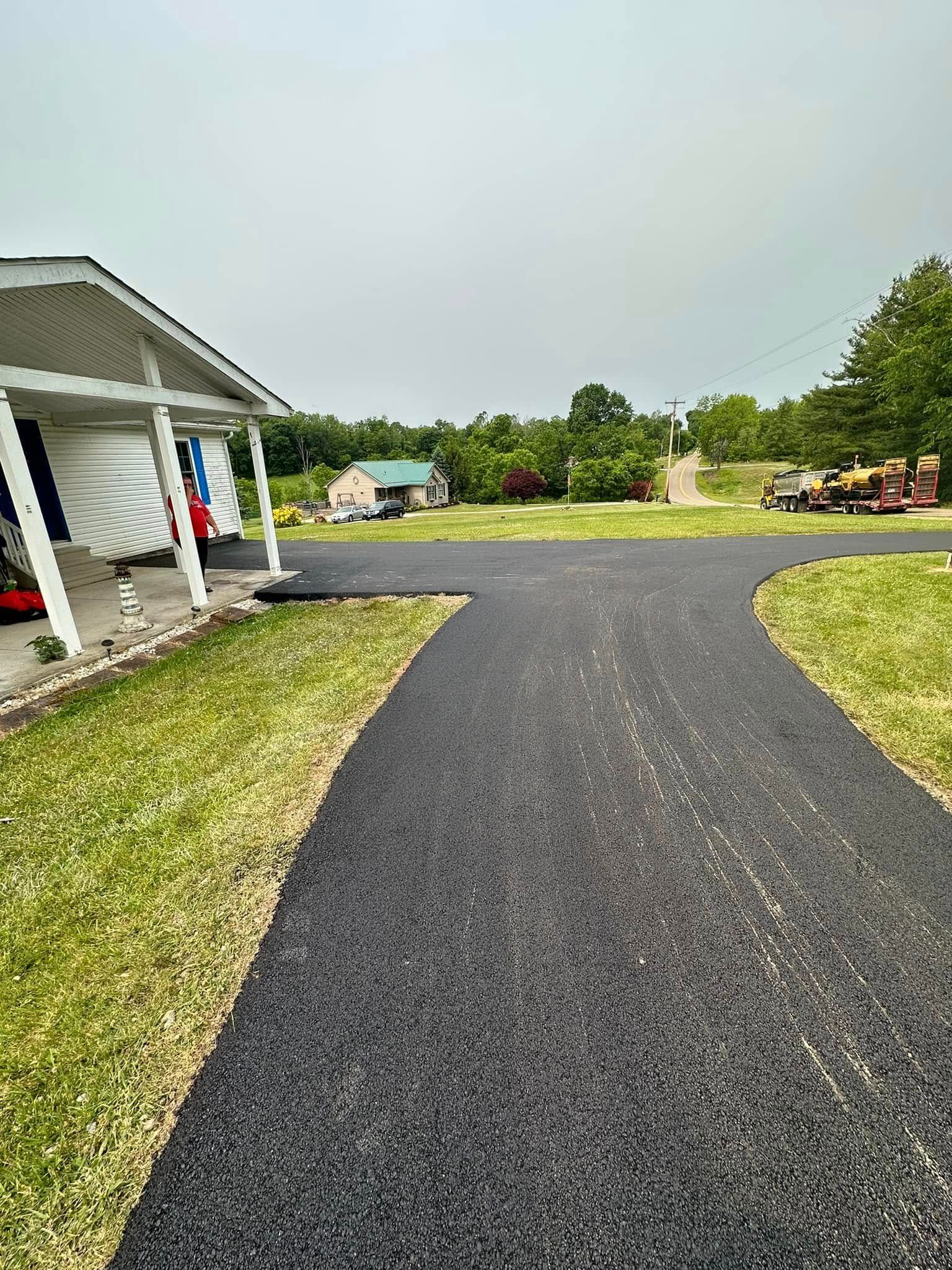 A freshly paved asphalt driveway extending from a house porch across a grassy lawn towards a distant outbuilding.
