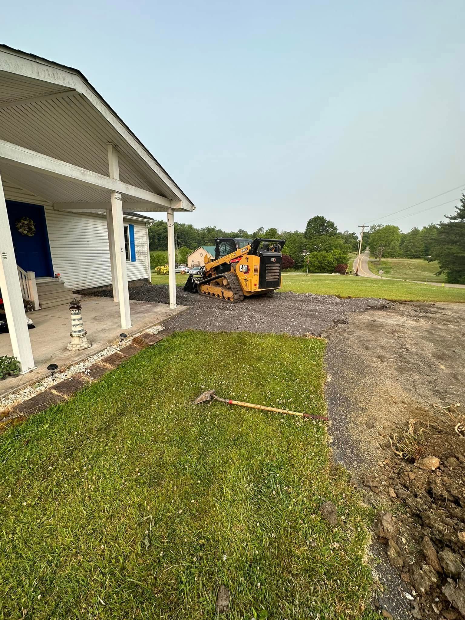 A yellow skid steer loader sits on a gravel driveway in front of a house with a porch and a grassy yard.
