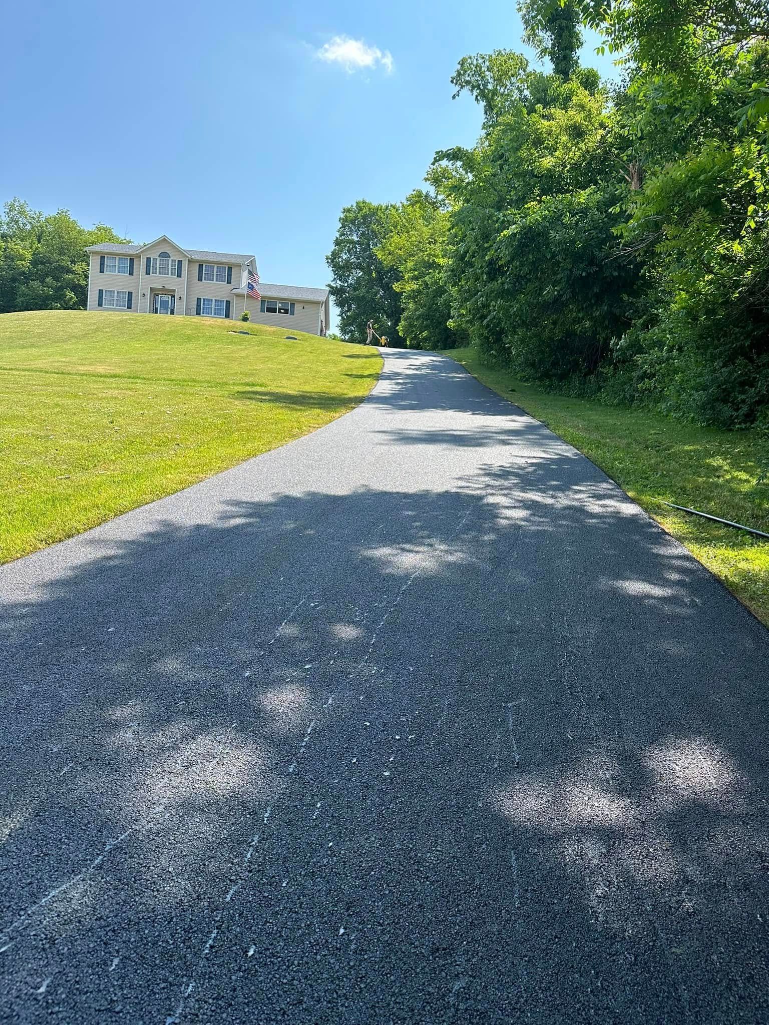 A long, paved driveway slopes upward toward a two-story house surrounded by green trees under a bright blue sky.