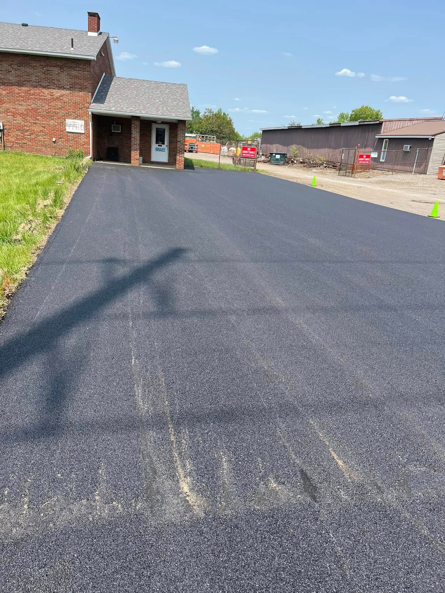 A freshly paved dark asphalt driveway leads to the entrance of a brick house under a clear blue sky.