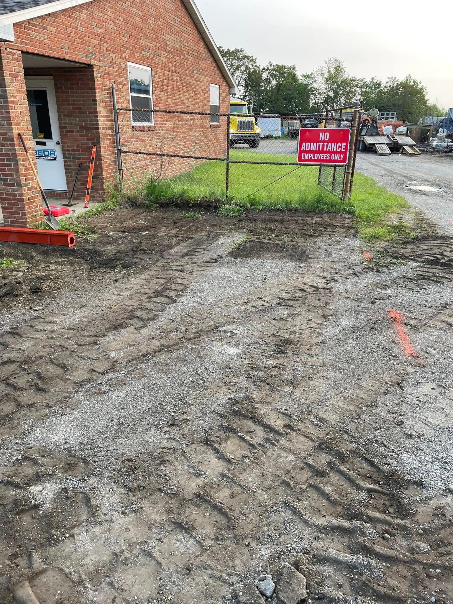 A red brick building exterior with a white door, adjacent to a dirt construction site with gravel, tire tracks, and a fence.