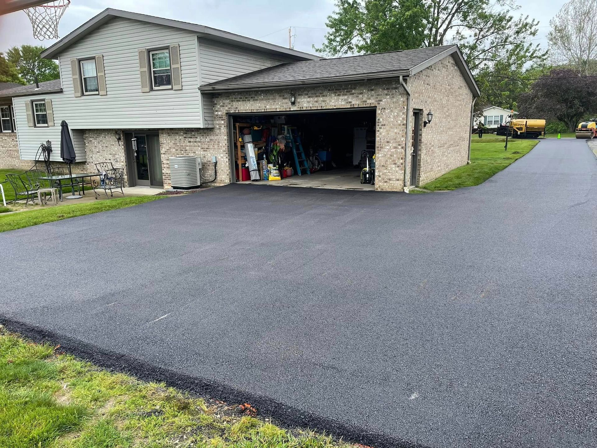 A newly paved asphalt driveway leads up to a two-story house with a brick garage, set against a green lawn.