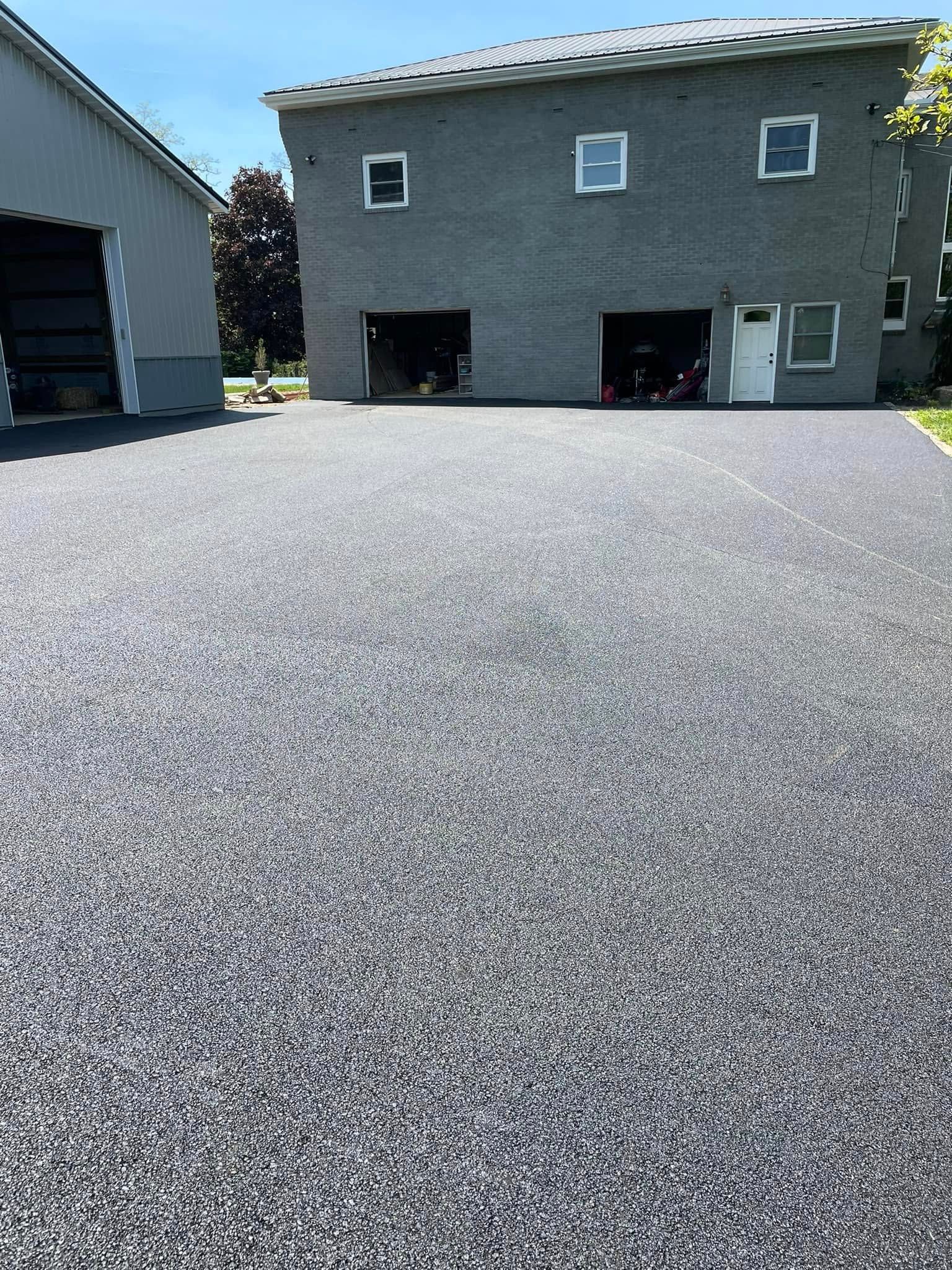 A freshly paved, dark gray gravel driveway in front of a gray brick building with three windows and two garage bays.