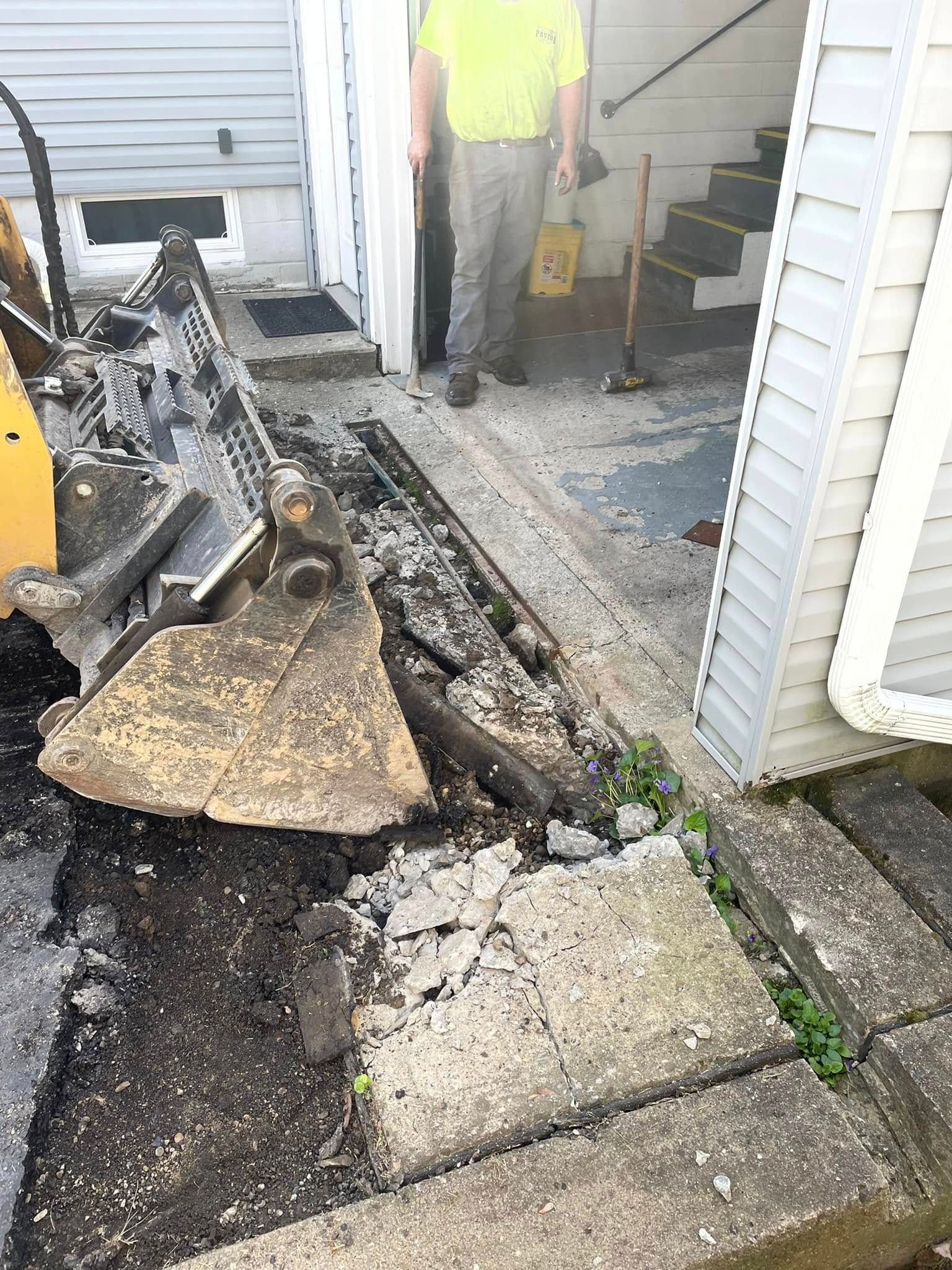 A person in a yellow shirt operates heavy machinery to excavate concrete debris next to a building entrance.