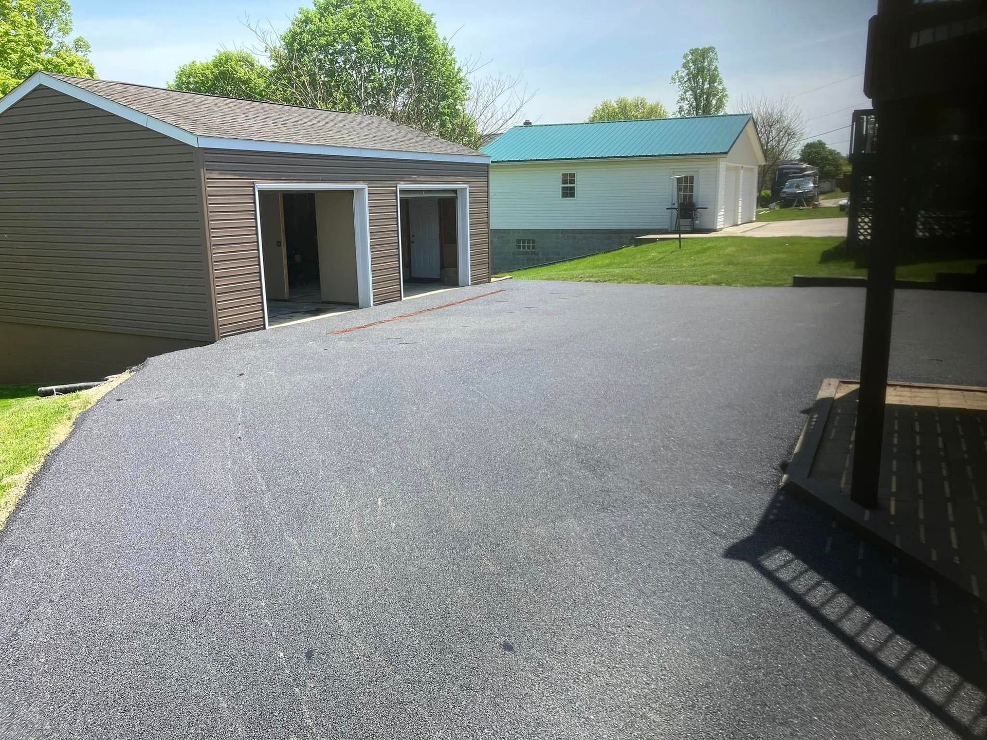 A freshly paved asphalt driveway leading to a two-car garage with a white building and green roof in the background.