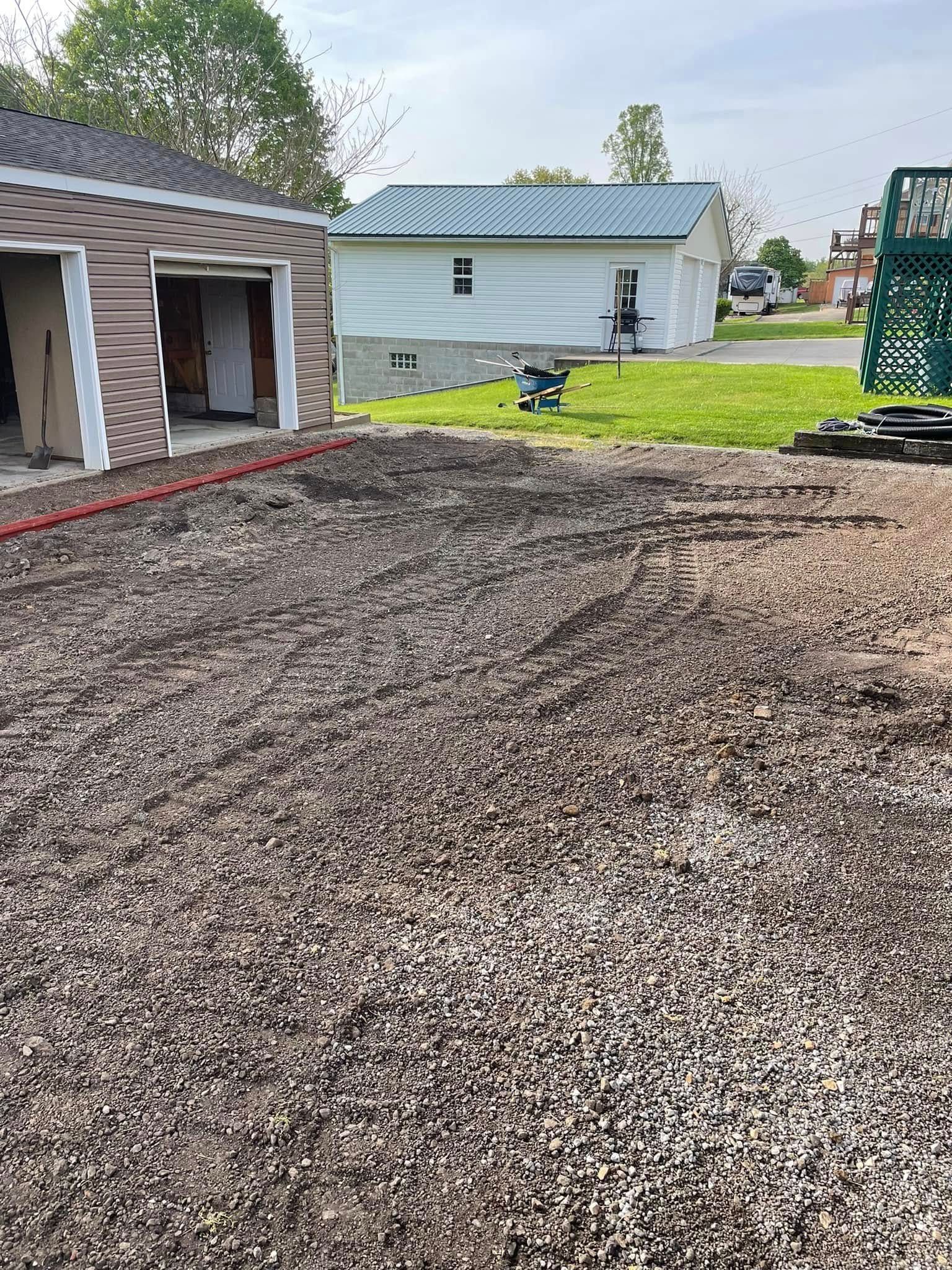 A gravel-filled yard under construction, bordered by a garage with brown siding and a white building with a green roof.