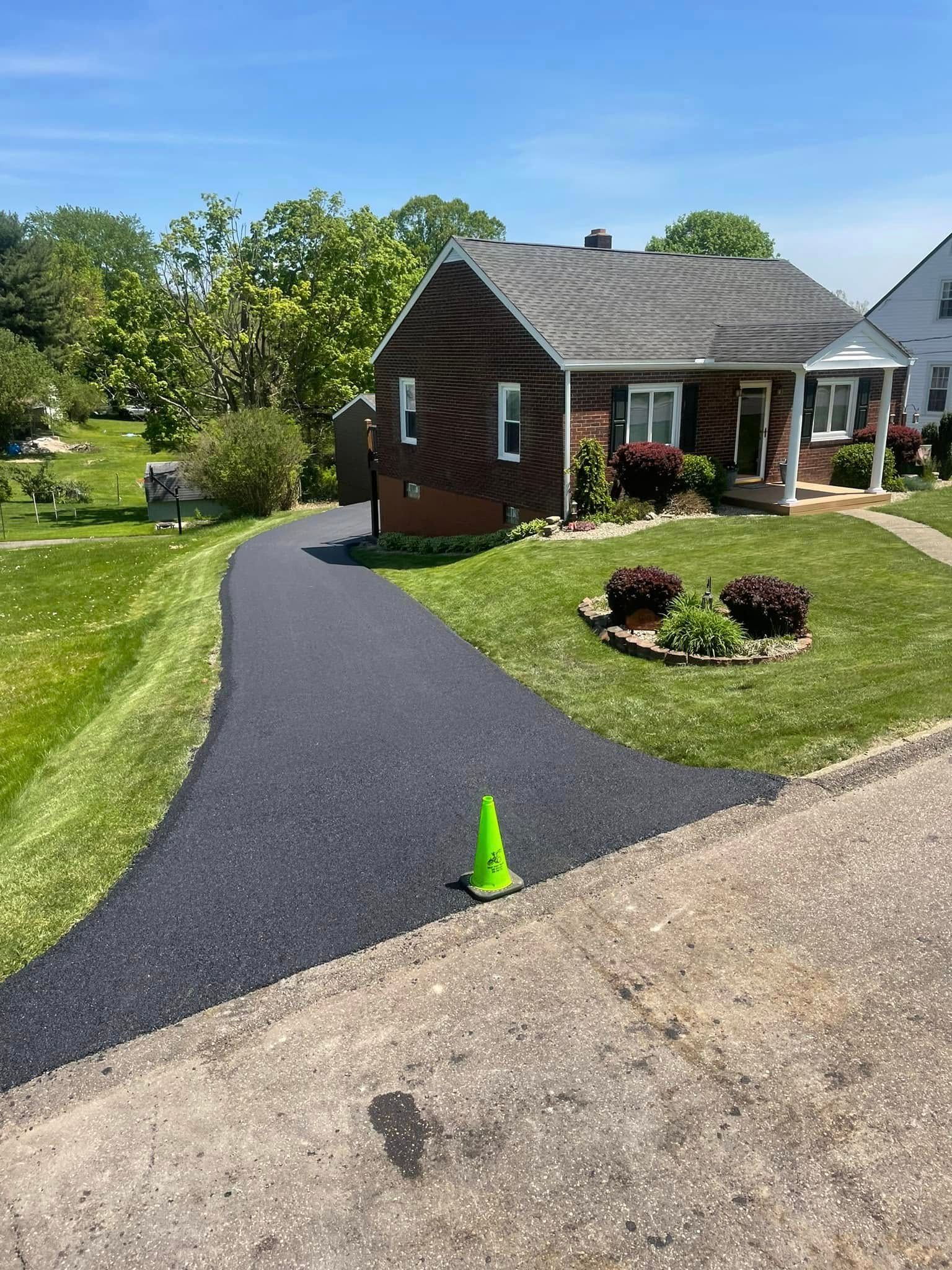 A newly paved, dark asphalt driveway leads to a small brown house on a sunny day, with a single green safety cone.