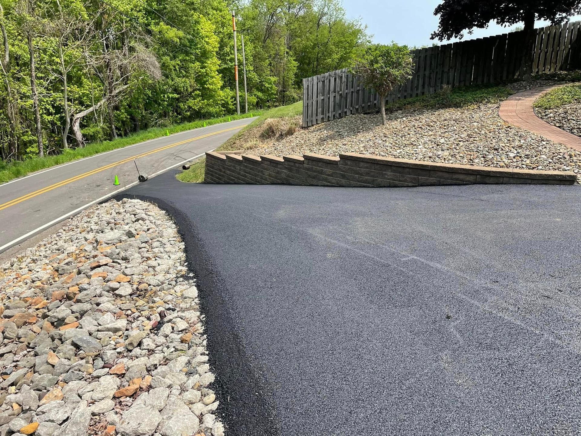 A paved driveway meets a rural road, bordered by sloped stone landscaping and a small stone retaining wall.