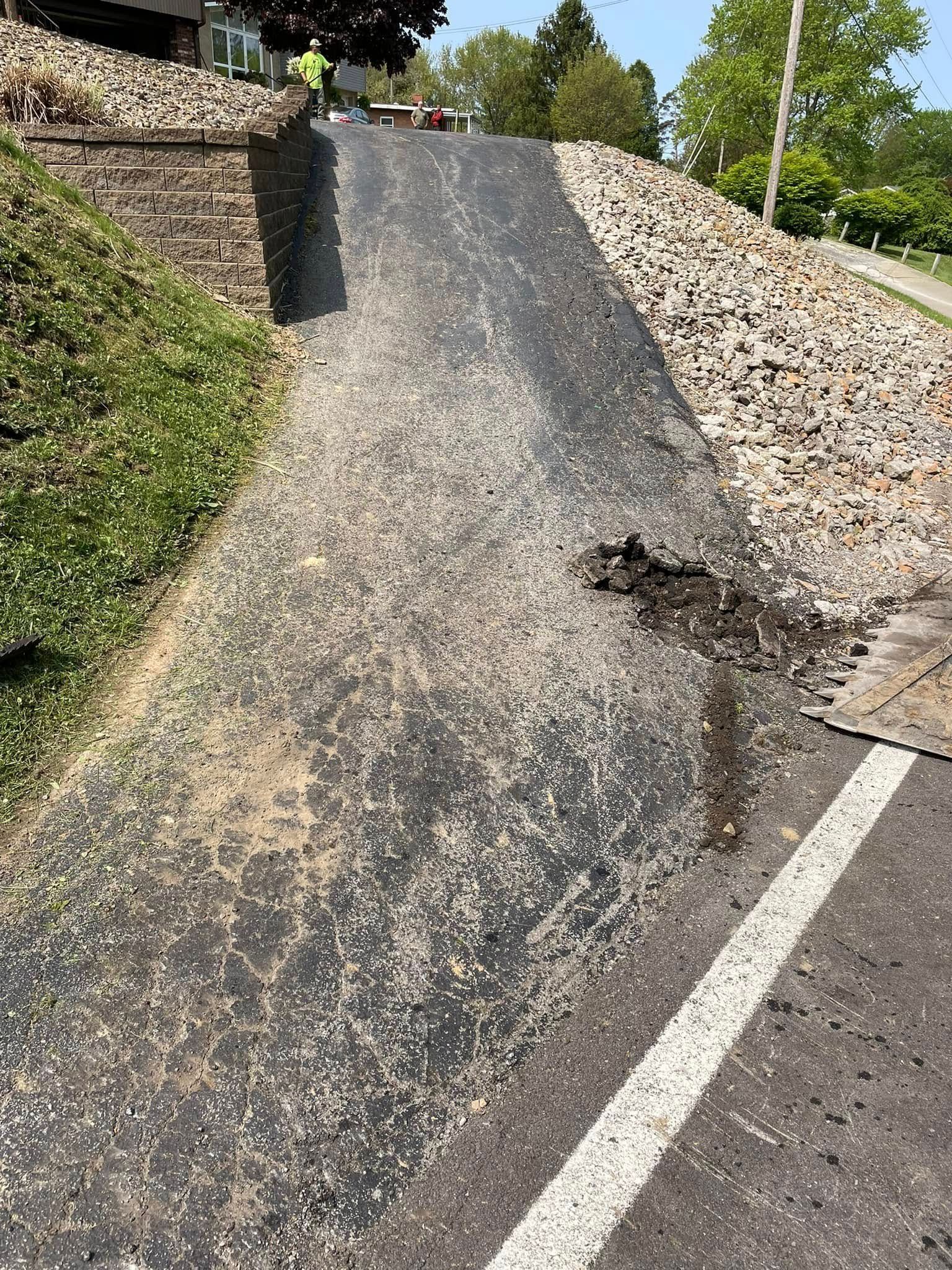 A steep, paved driveway sloping upward between a grassy embankment on the left and a large pile of rocks on the right.