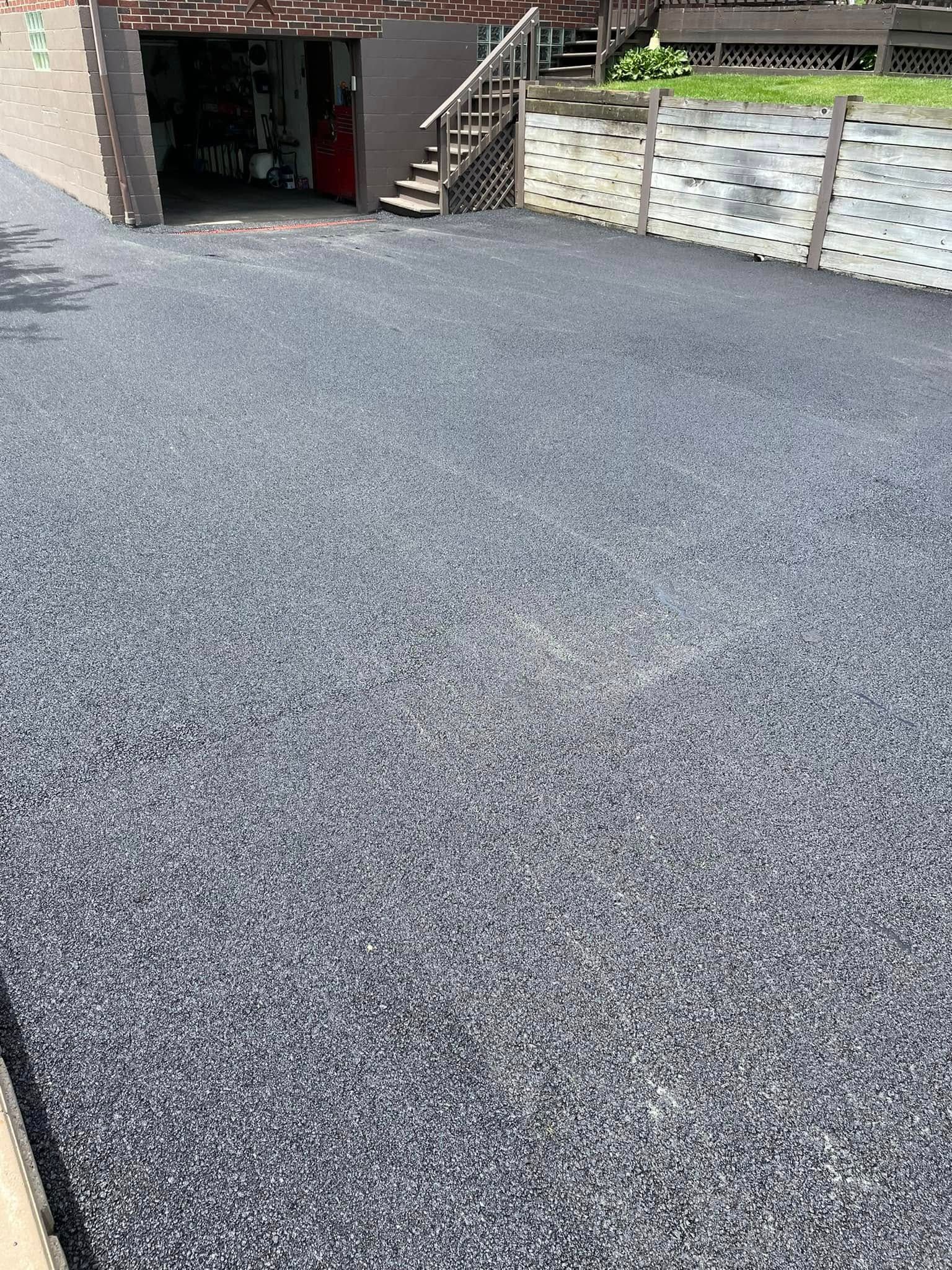 A freshly paved dark gravel driveway leads toward a garage entrance, featuring a small staircase and a retaining wall.