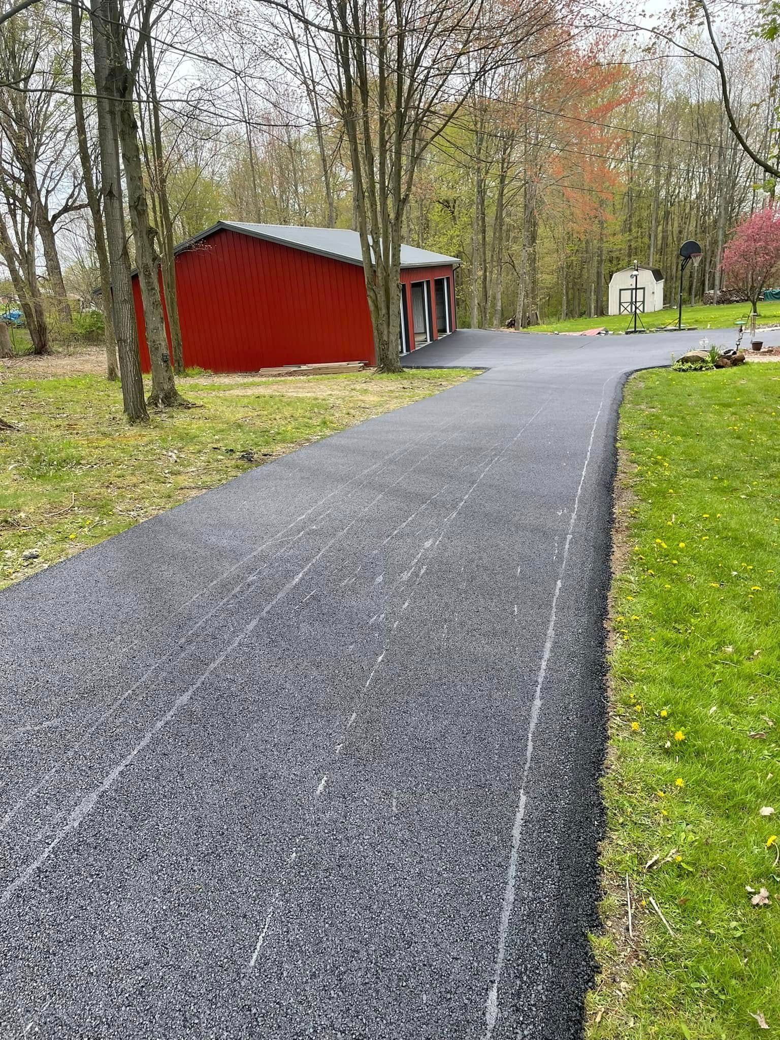 A newly paved asphalt driveway leads to a small red barn structure surrounded by trees and grass in a park-like setting.