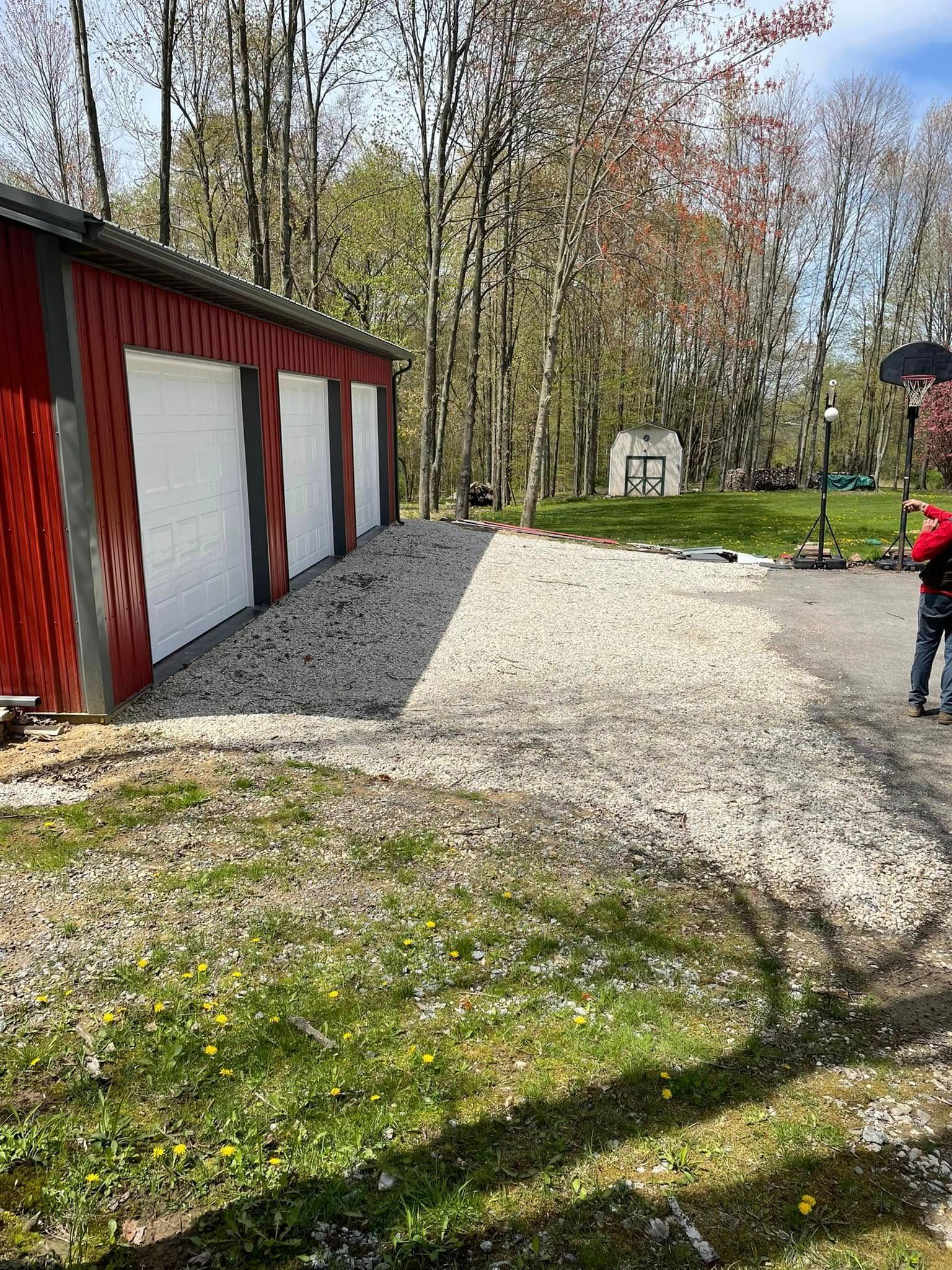 A red three-bay garage with white doors sits beside a gravel driveway, with trees and a small white shed in the background.