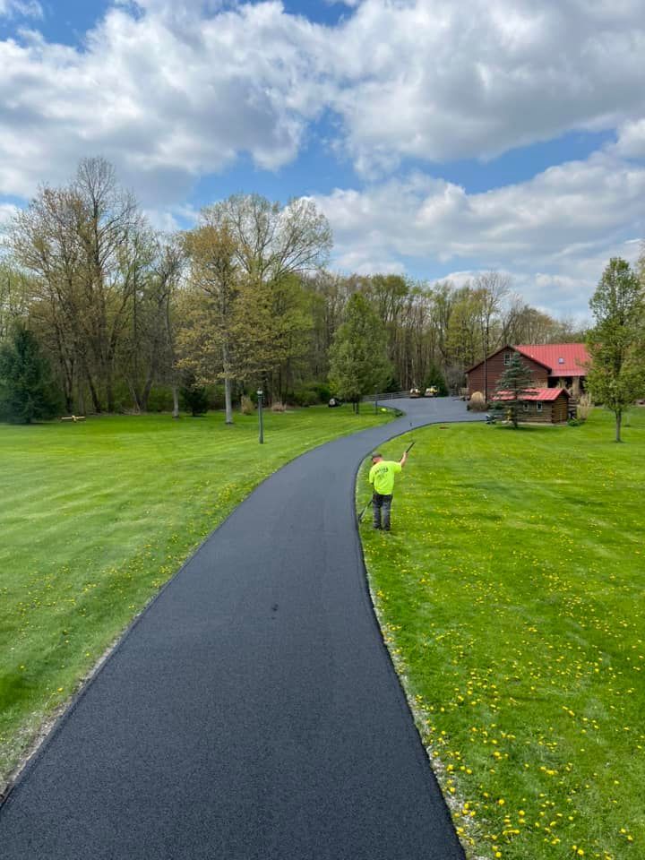 A person in a neon yellow shirt walks along a newly paved black driveway leading to a red-roofed building on a grassy lot.