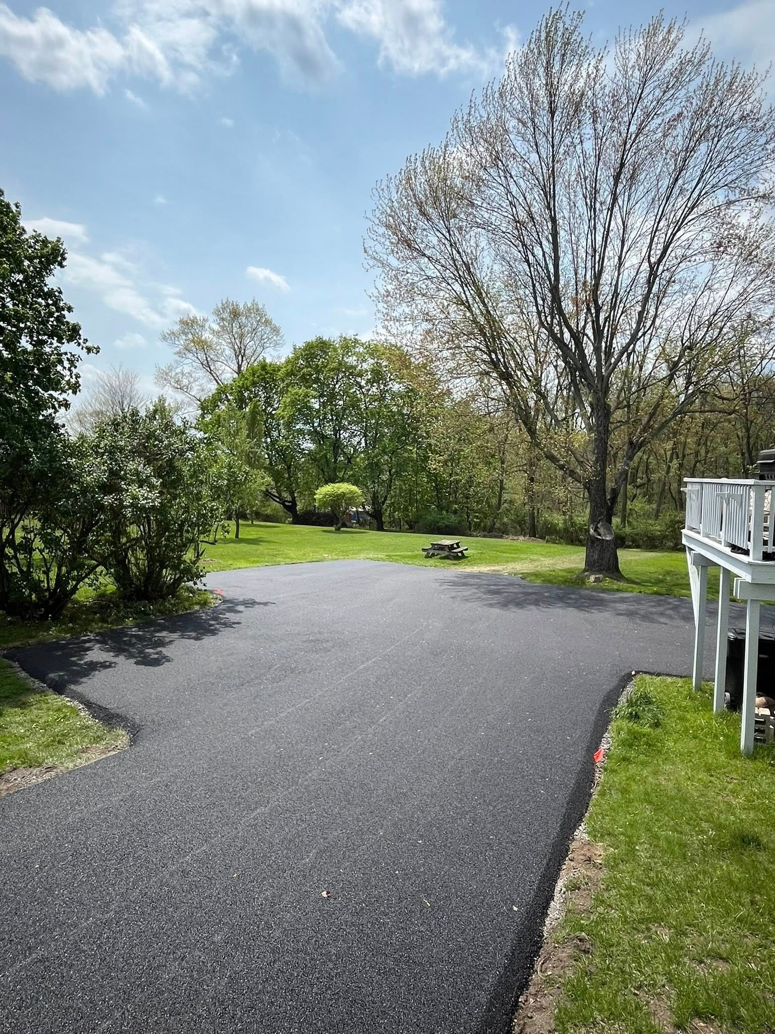 A wide view of a freshly paved black asphalt driveway leading into a grassy backyard with trees and a wooden deck.