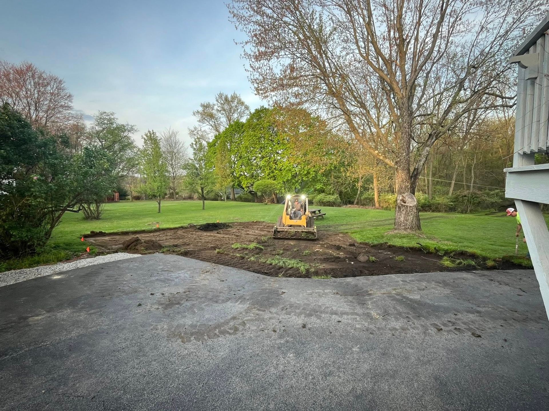 A yellow skid steer sits on a freshly cleared patch of dirt at the edge of a paved driveway in a grassy backyard.