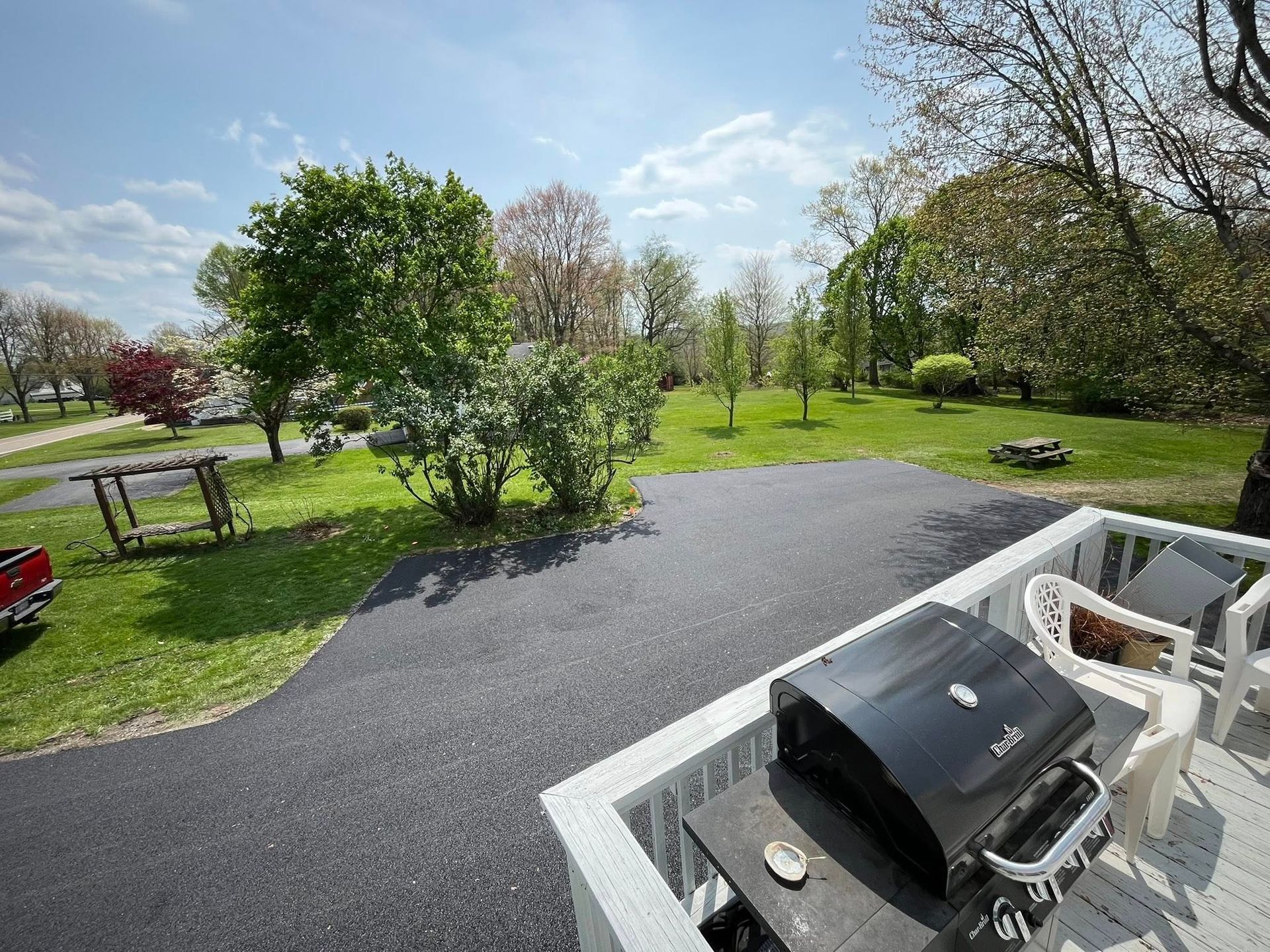 A black grill sits on a white deck overlooking a paved driveway, a grassy yard, and trees under a blue sky.