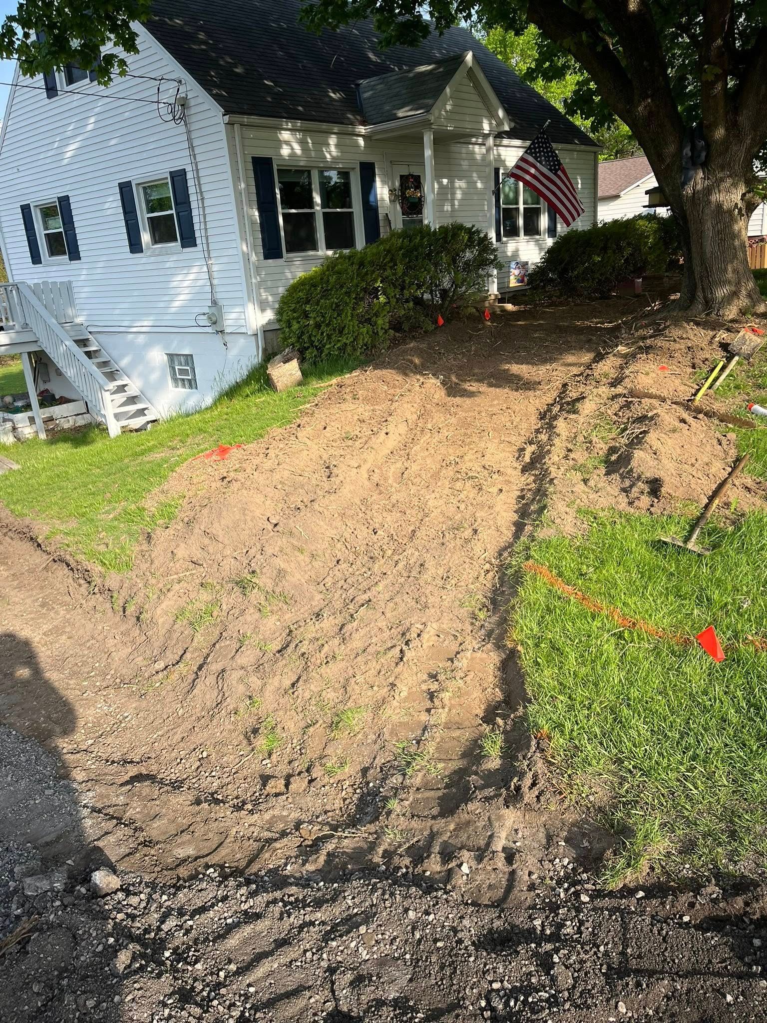 A white house with a dark roof and blue shutters sits behind a freshly excavated dirt slope with a nearby orange marker.