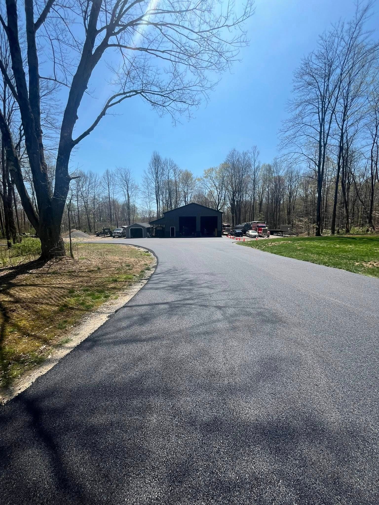 A freshly paved asphalt driveway leads toward a dark grey detached garage surrounded by trees under a bright blue sky.